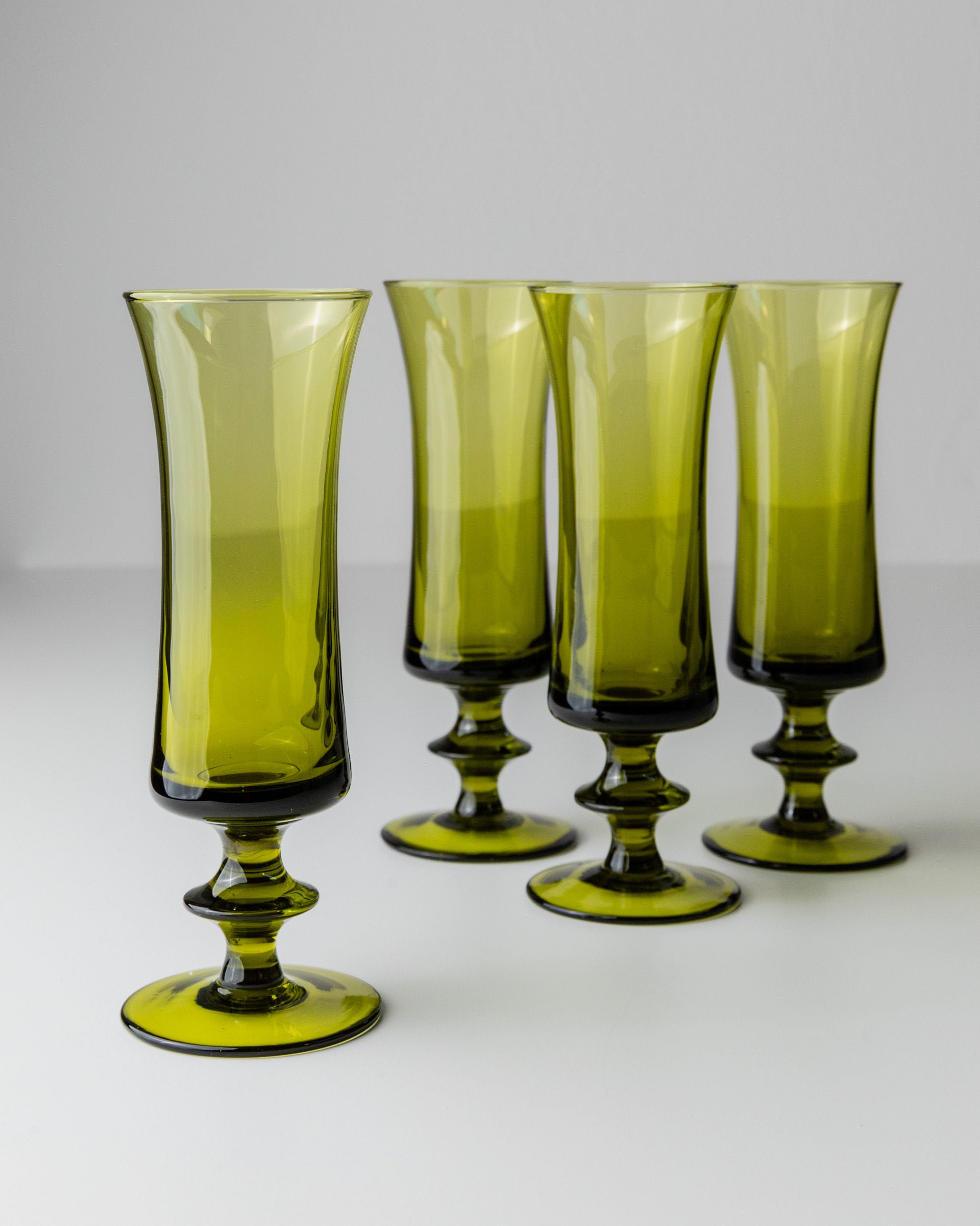 The Bottega Jacobs 4 Murano Glasses in Green 70s, with their wide flared rims and ornate stems, showcase a retro chic style on a neutral surface.