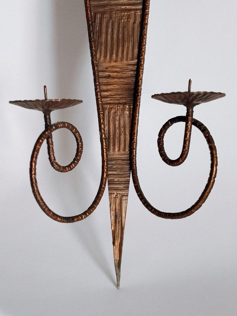 The Huerto Metalwork Wall Candleholder features two holders with ornate spiral arms and artisan hammered detailing on its pointed central backplate, showcased against a plain light background.