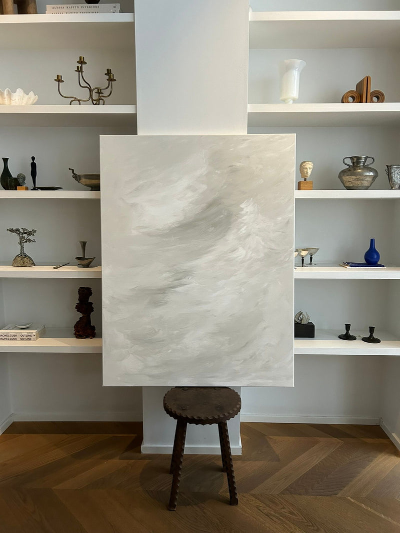 The "Calm in Motion" painting by Mattias Storm, a mixed media on canvas in gray and white, is positioned on a stool before built-in shelves with decorative vases, sculptures, and candlesticks against a light-colored wall.