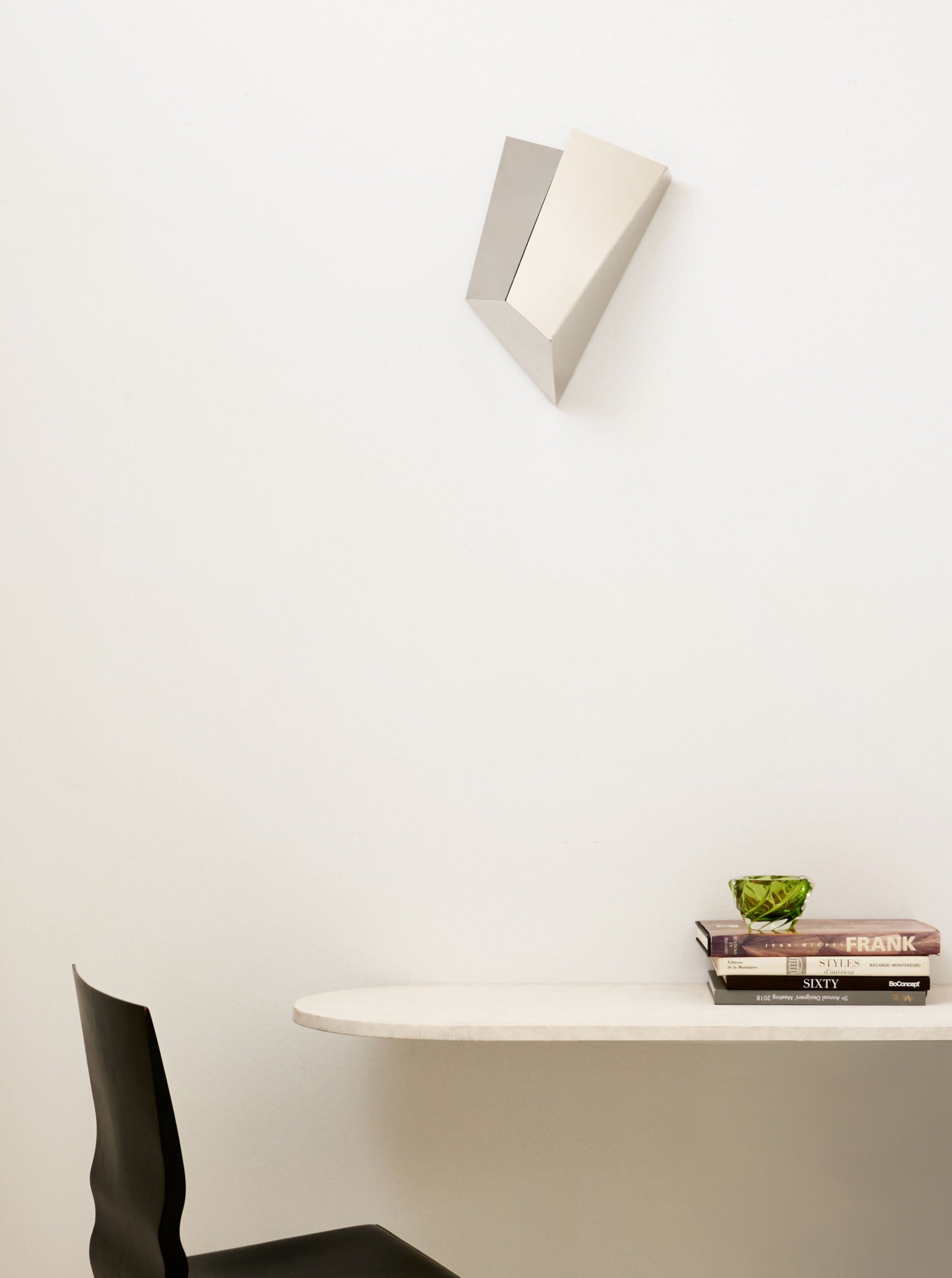 Minimalistic interior featuring a white wall with a Mariza Galani Katoptris Nickel wall lamp above a sleek shelf holding books and a decorative green plant, with a black chair adjacent to it.