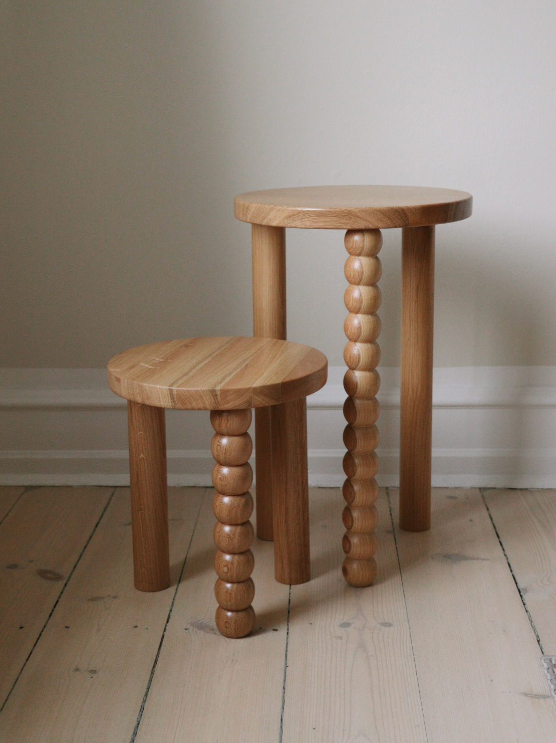 Against a neutral wall on a light wooden floor, two wooden stools with distinctive peg-like legs are inspired by Maitoinen Home's Piloti Side Table in Oak. The taller stool has one decorative turned wood leg and the shorter has all legs in this style, showcasing simple Euclidean geometry.