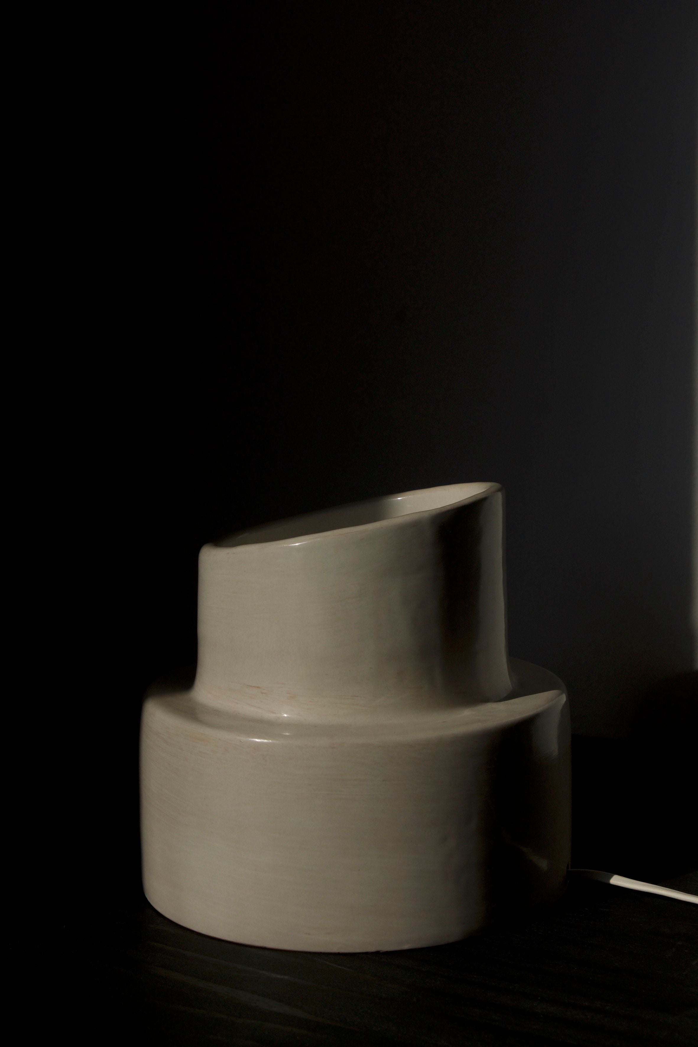 A beige, modern ceramic vase with an asymmetrical top sits on a dark surface, softly illuminated by COSEINCORSO's "Pillar" Table Lamp on the right, all set against a dark background.