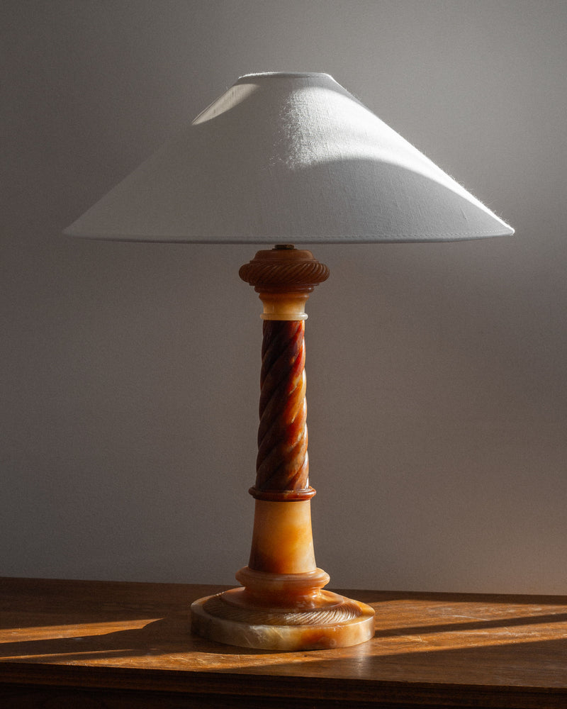 The Antique Alabaster Table Lamp, C. 1900 by Spigel exudes a classic and timeless aesthetic with its hand-carved alabaster base featuring a twisted column design. Resting on a wooden surface, its wide off-white lampshade is softly illuminated by natural light from the left, casting enchanting shadows on the wall behind it.