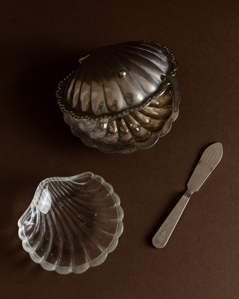 A Silver-plated Butter/Caviar Dish from the 1950s by Bottega Jacobs, designed in the shape of a seashell with a lid, accompanied by a small glass dish resembling an Italian shell and a metal spreader, is elegantly displayed on a dark brown surface.