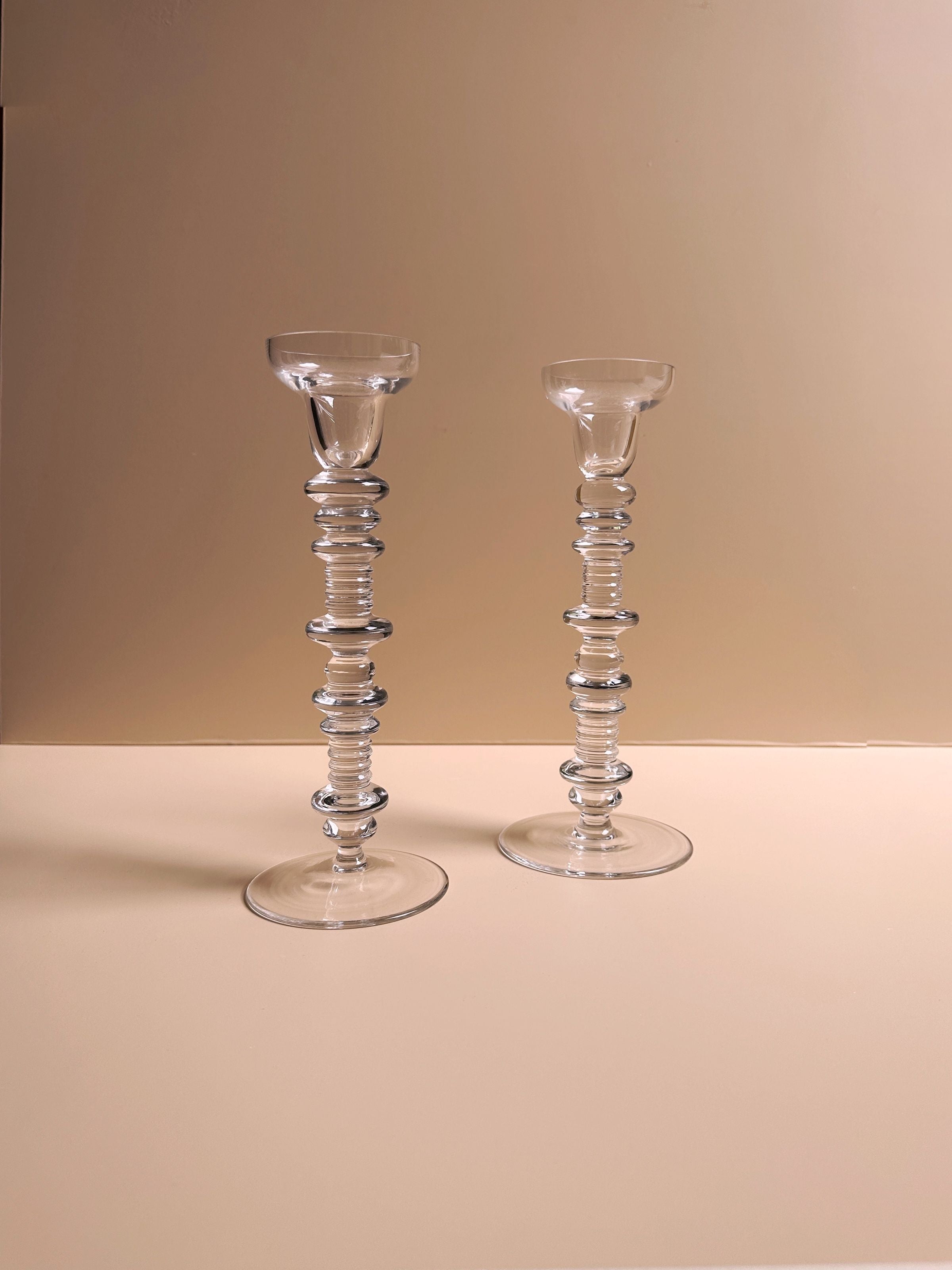 The Set of 2 Glass Candleholders by Peill & Putzler from Dodo Vintage showcases a mid-century design with stacked circular patterns and slight height variations. Their modern, transparent look beautifully complements the beige surface and matching wall, embodying the timeless elegance synonymous with Peill & Putzler.