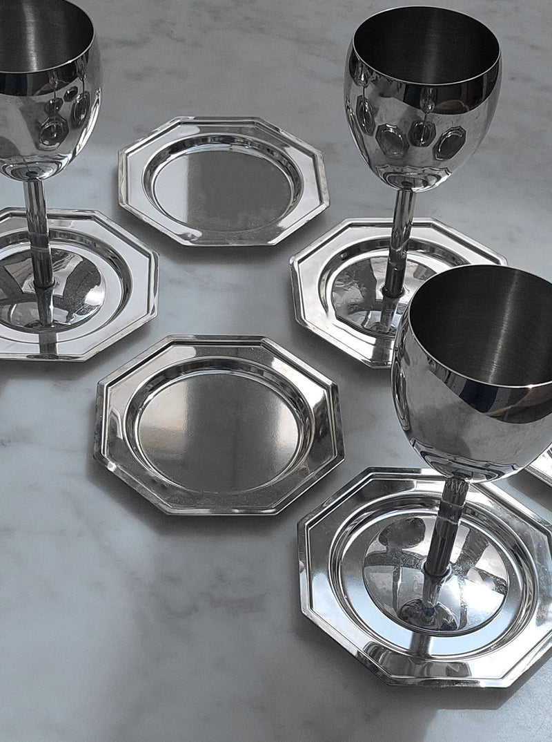 A set of four polished silver wine goblets paired with a Set Of 6 Coasters by Les Objoies, arranged artistically on a marble tabletop. The reflective surfaces of the goblets and the octagonal glass coasters glisten under the ambient light, showcasing a modern design crafted from durable materials.