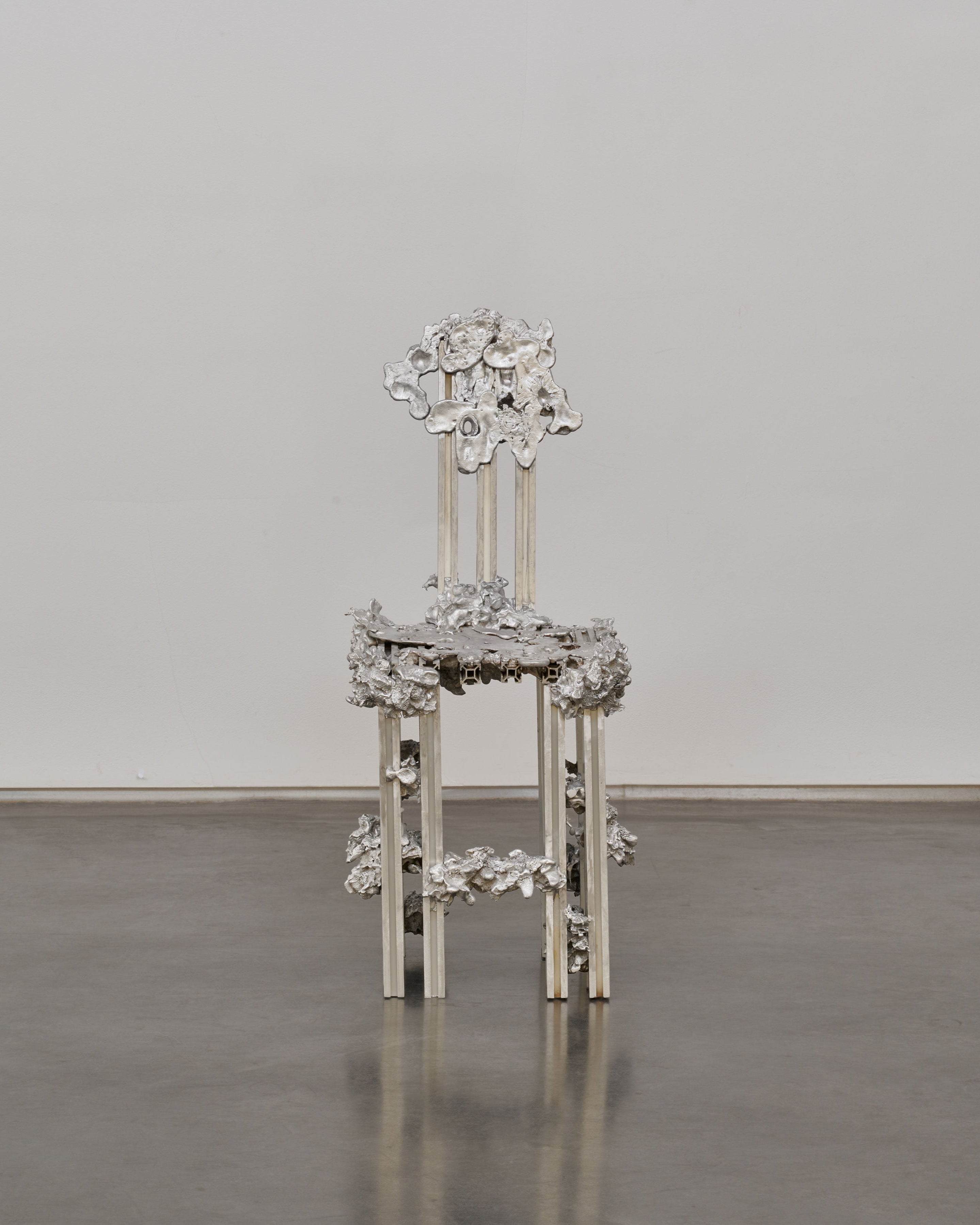 The 'Graft' Chair in Cast Aluminum by Sheyang Li features intricate, decorative patterns and an ornate silver design that blends industrial precision with organic growth, standing out against a plain white wall and polished concrete floor.