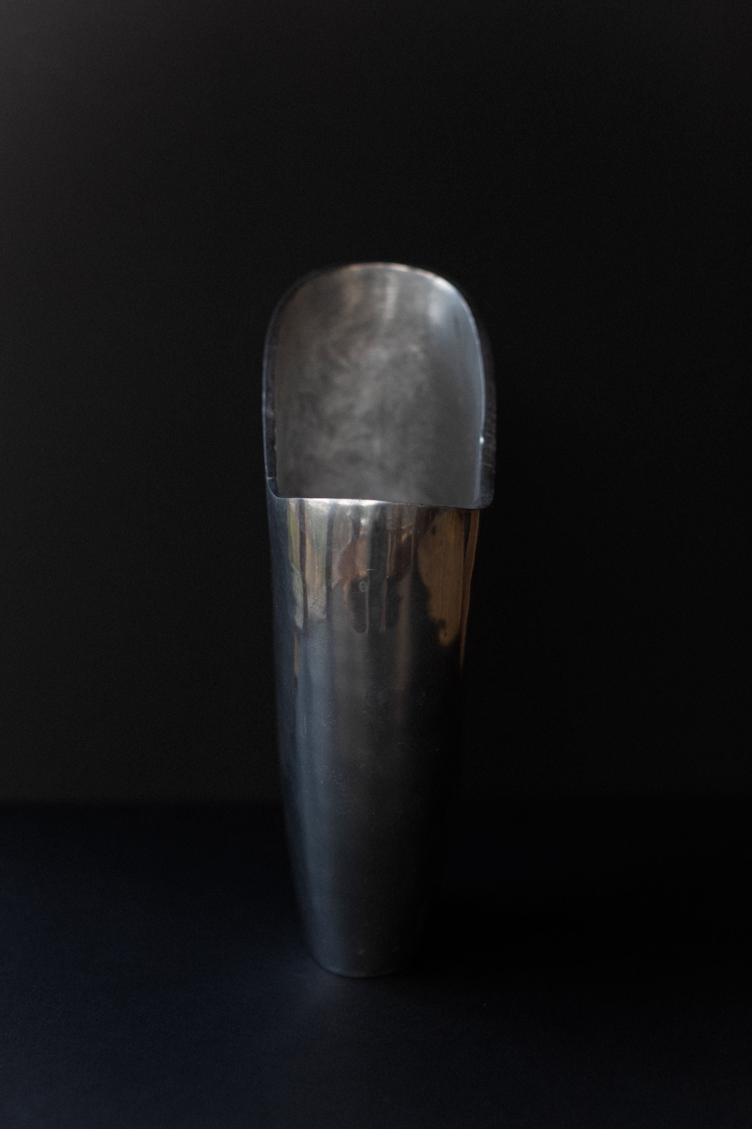 An Aluminum Cast Vase from Out For Lunch stands upright against a dark background, reflecting light on its curved surface. With a smooth, shiny finish and wide, rounded shape, it exudes an industrial aesthetic akin to a sculptural presence.