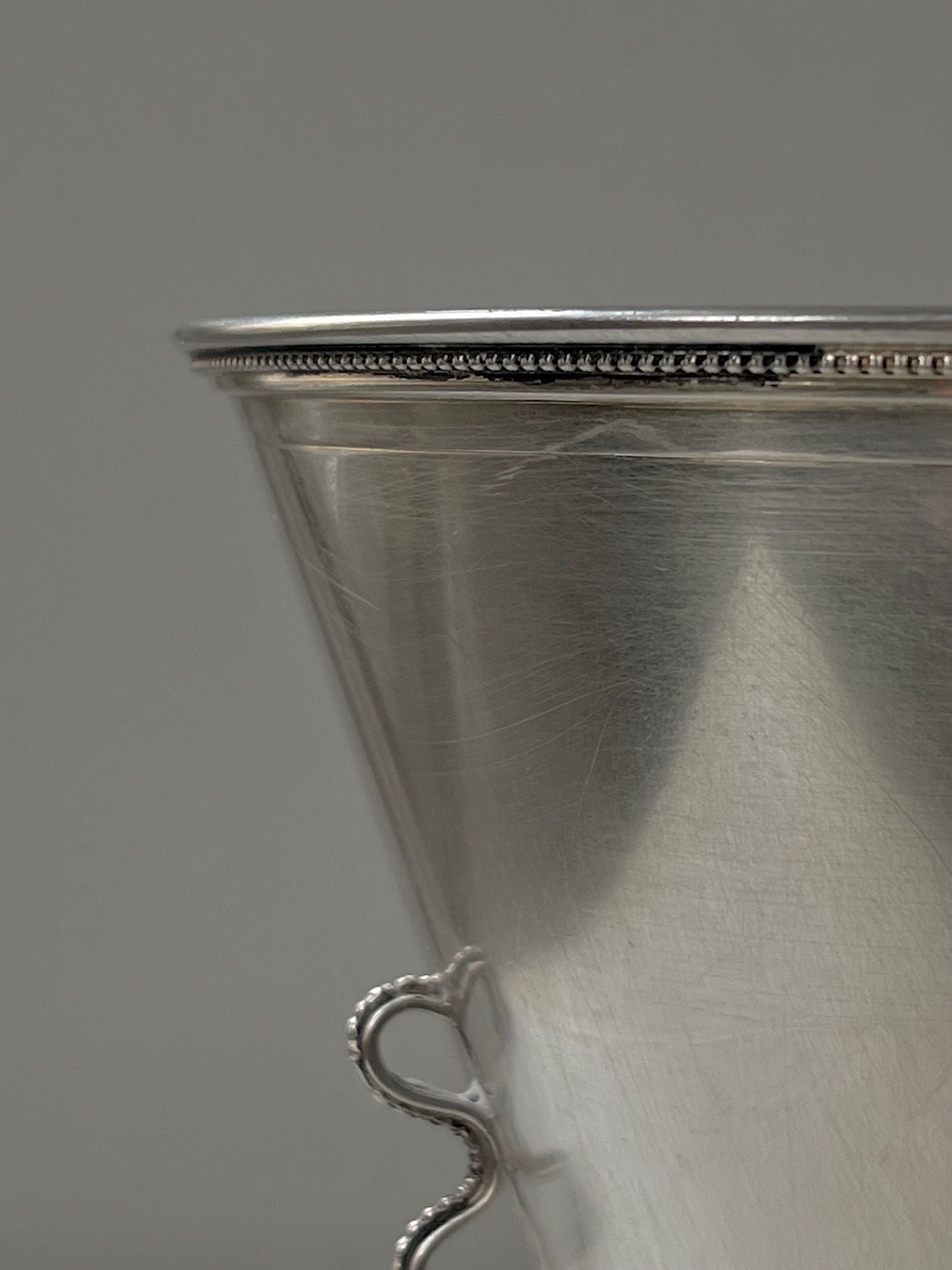 Close-up of a hand-worked 1930s Silver Vase by Médecine, showcasing detailed craftsmanship. The upper rim features a beaded design, with a partially visible decorative handle. Its slightly textured surface softly reflects light against a plain background, reminiscent of the elegance found in Stockholm's artisan markets.