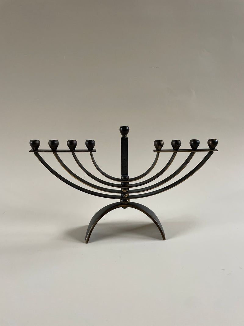The Maud Vaughan Menorah Candelabra features a vintage brass design with nine candle holders arranged in a semicircular fashion, resting on a sleek curved base against a neutral backdrop. It gracefully holds slender taper candles, lending an enduring charm to its classic silhouette.