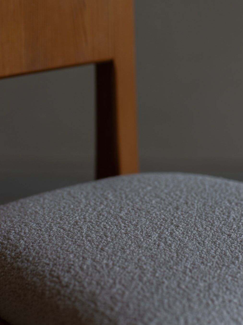 Close-up of a Bicci de' Medici Göran Malmvall - Model 553 chair with a textured gray upholstered seat against a neutral dark background, highlighting the contrasting textures of the wood and fabric, exemplifying exquisite Swedish furniture design.