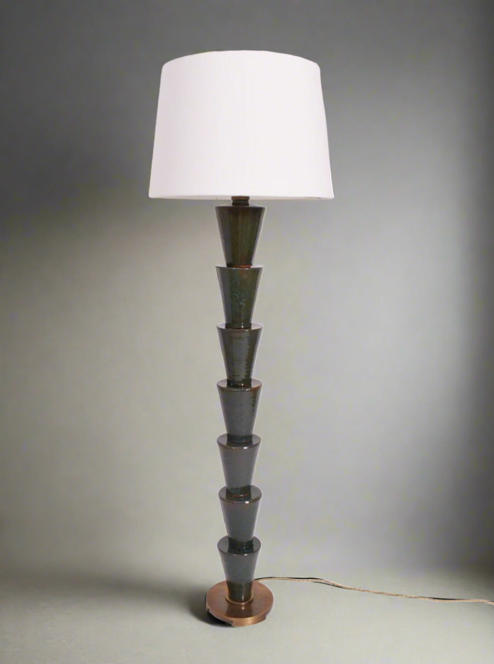 A tall, modern "Nizwa" Floor Lamp by Barracuda Interiors featuring a white lampshade and a stacked, cone-shaped, stoneware glazed base. The lamp is proudly made in Portugal, placed against a white background with a visible power cord running along the floor.