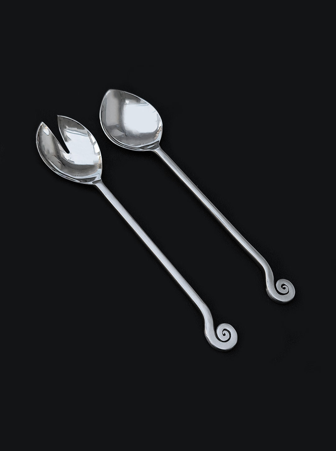The Les Objoies Spiral Salad Servers feature two metal utensils—a solid spoon and a forked server—with long handles and spiral ends, each in polished brass for a decorative touch against a dark background.