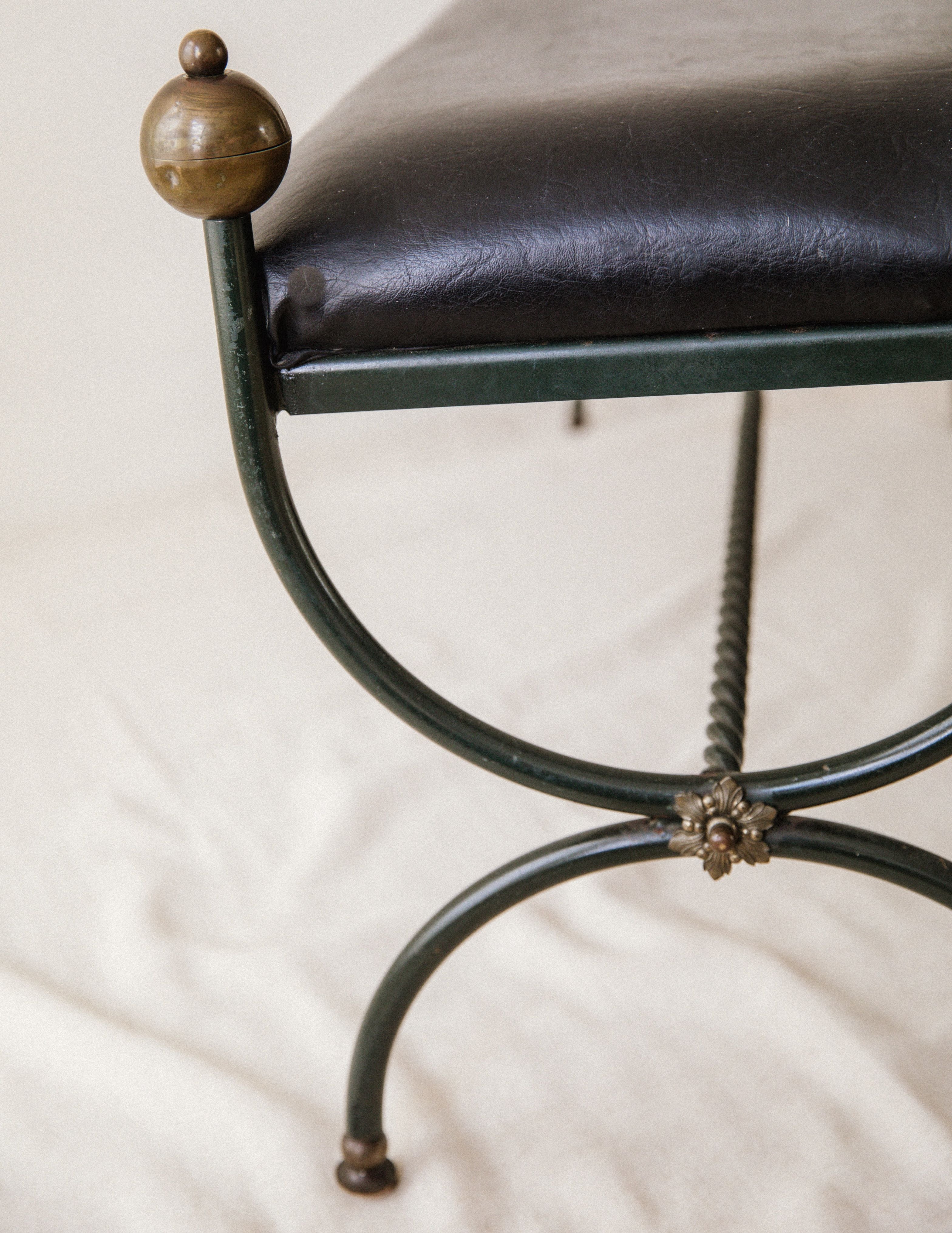 Close-up of the Arcā Collections Mid-Century Wrought Iron Bench, featuring a black leather cushion, green curved legs, decorative brass details, and twisted metal supports, shown resting on a light fabric surface.