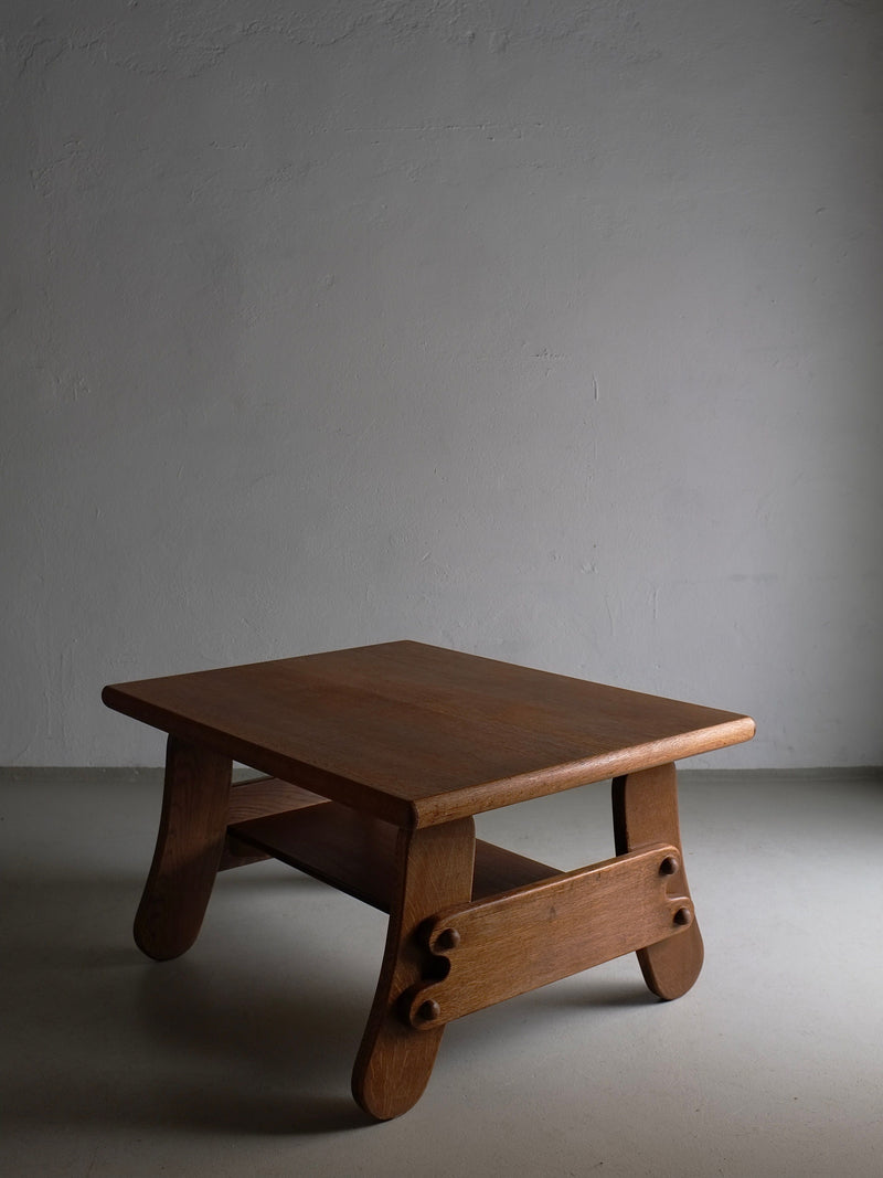 A Brutalist Oak Coffee Table | Netherlands | 1970s by Veter Vintage with curved legs, a lower shelf, and a rectangular top is placed against a plain grey wall. The table's design features simple, smooth lines and a natural wood finish reminiscent of 1970s design. The lighting casts soft shadows, emphasizing its texture and shape.