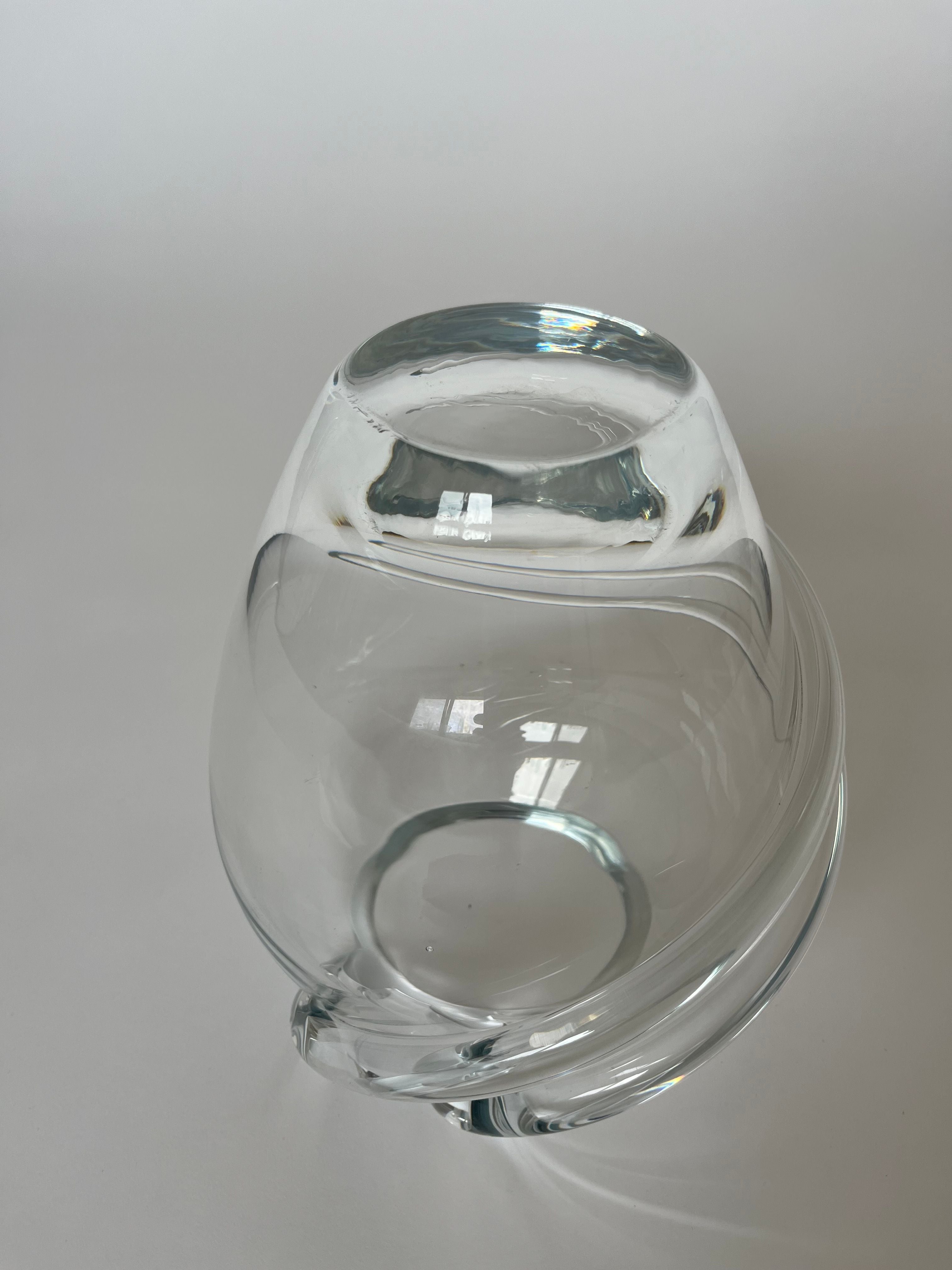 A Vintage Crystal Vase by septembre studios, showcasing a modern, rounded design, is placed on a white surface. This clear glass vase features a smooth, reflective finish and a wide open top, complemented by delicate swirl details. The background remains plain and minimalistic.