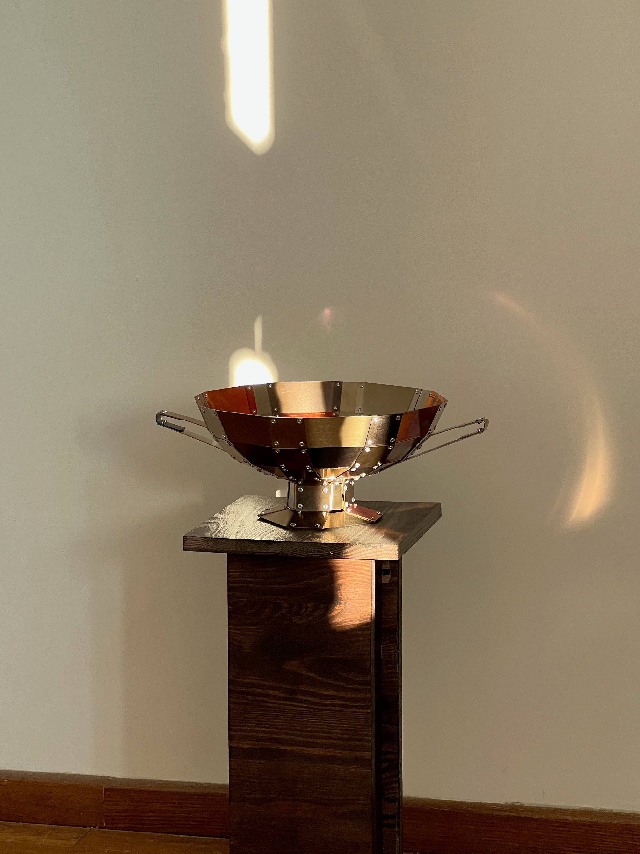 The Theo Galliakis Kylix, a metallic cup with handles, sits on a dark wooden pedestal against a plain wall, bathed in warm natural light that casts soft Greek-inspired reflections and shadows.