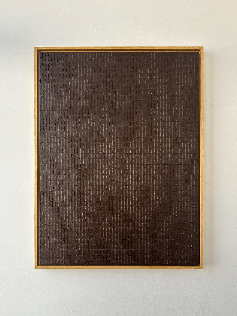 The "Burnt Umber - Oil on Canvas" by Roi Elmaliah is elegantly displayed in Amsterdam. This rectangular piece boasts a dark brown, textured surface with a grid pattern. It is crafted with oil on canvas and framed in a simple wooden border, set against a plain light-colored wall to enhance its sophistication.