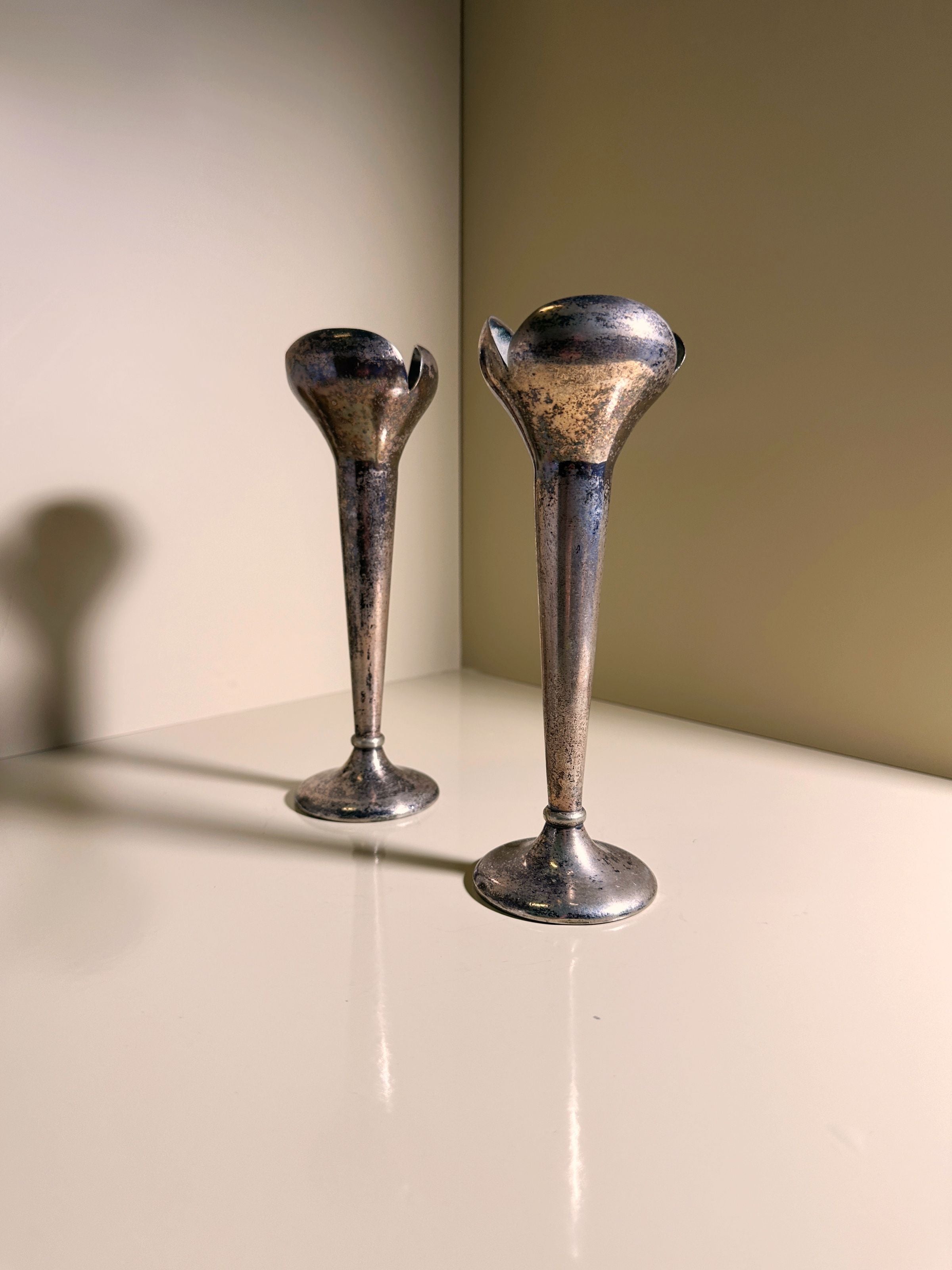 Displayed on a light surface, the Set of 2 Candlesticks by Gio Ponti for Calderoni from the 1930s, offered by Dodo Vintage, features a tarnished metal finish with flared tops reminiscent of trumpet shapes. The neutral background casts soft shadows from the candlesticks.