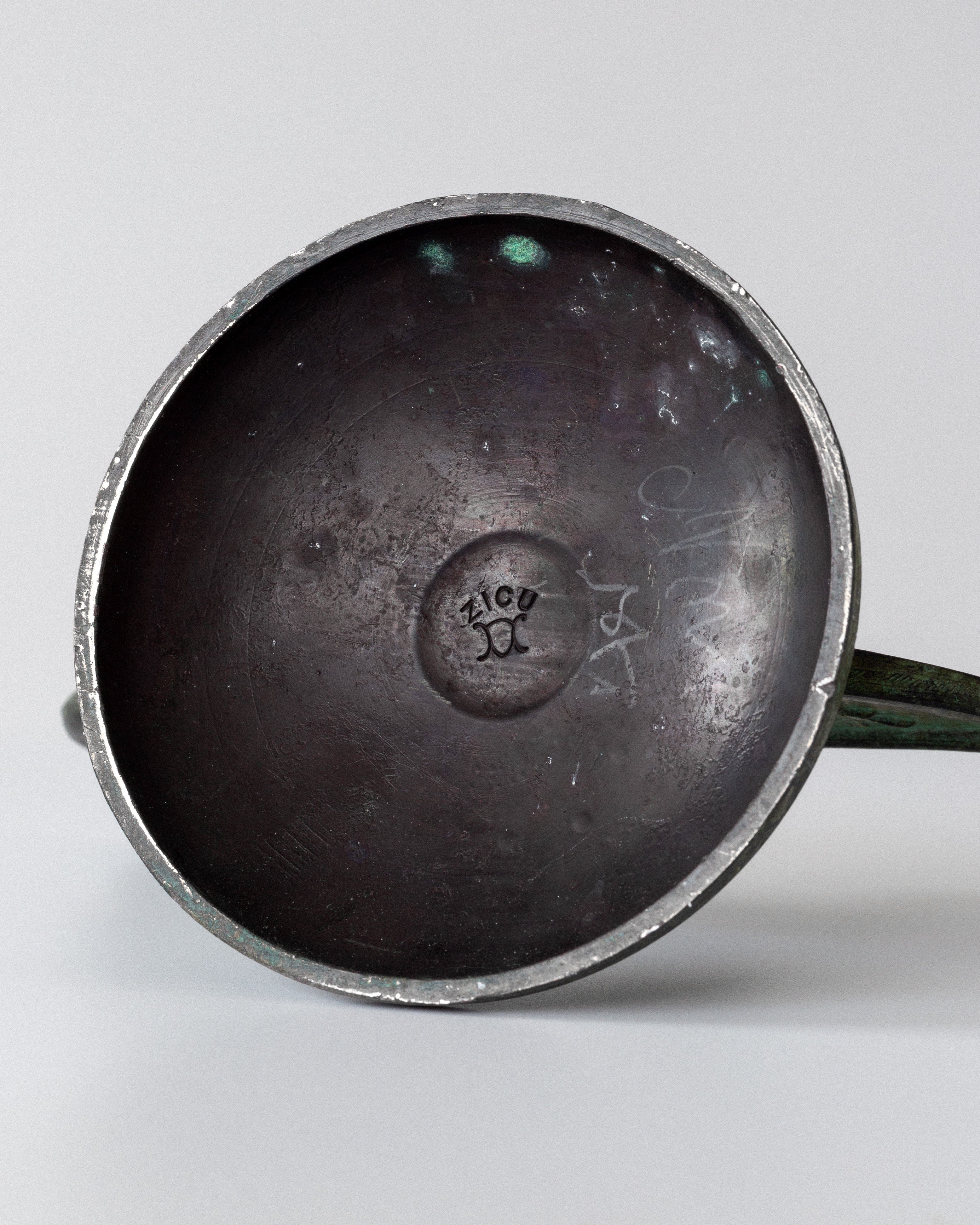 A close-up of a round, metallic underside reveals a patinated bronze finish with weathered and dark features. The center is stamped "Zico," while a thin rod extends from it, resembling the design of Spigel's Pair of Art Deco Bronze Candleholders.