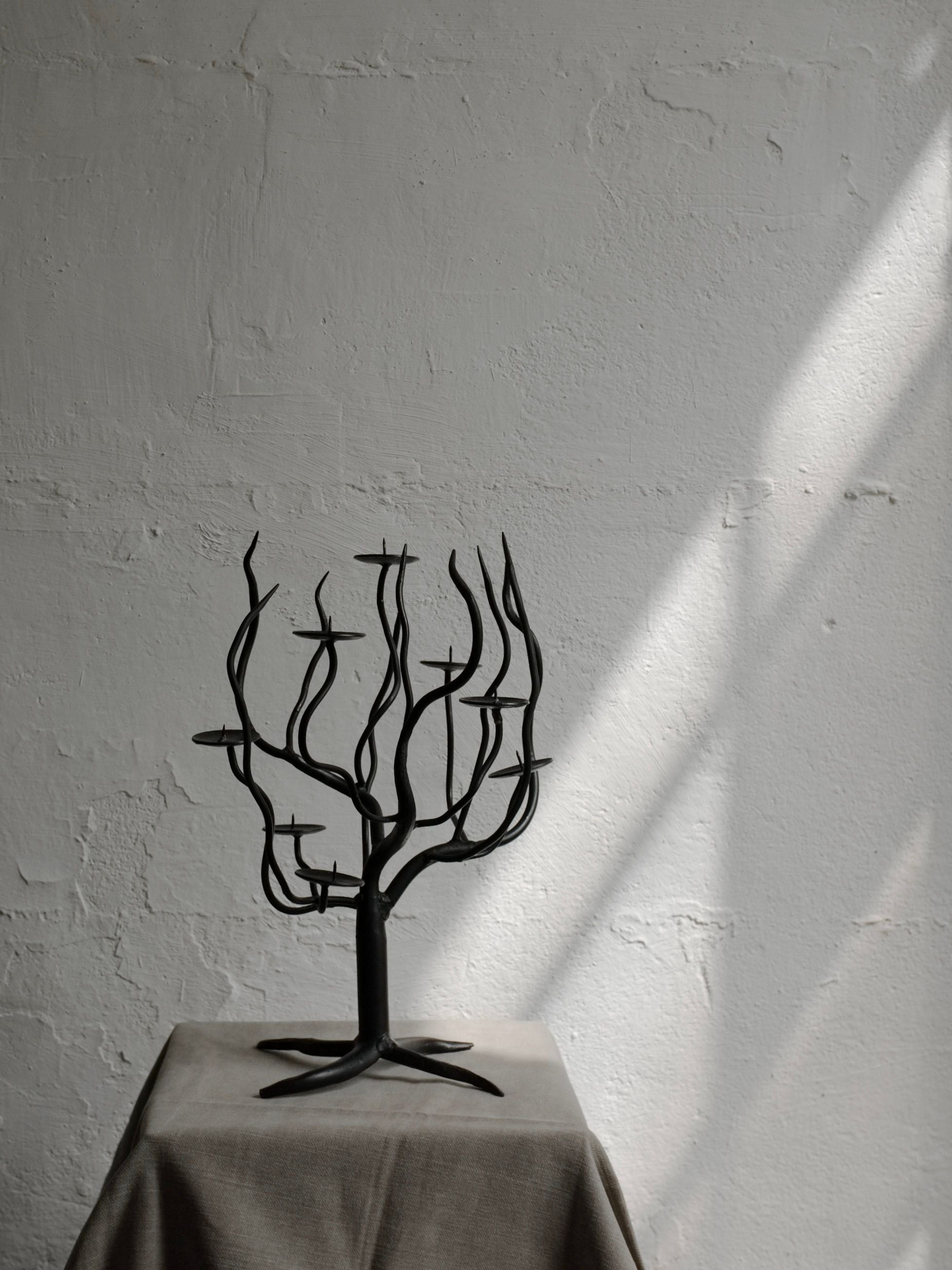 The Hand-forged Metal Sculptural Candle Holder by Veter Vintage, with its minimalist black metal design reminiscent of a leafless tree from the 1960s, stands prominently on a gray cloth-covered pedestal. The background features a textured white wall, illuminated by sunlight streaming in from the right, casting gentle shadows.