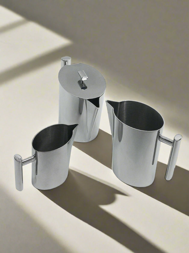 Three stainless steel kitchen containers with handles from the Vintage Coffee Set by Les Objoies are placed on a light-colored surface. This elegant coffee or tea set varies in size; one has a lid with a small handle, while the other two are open-top pitchers without lids. The containers cast distinct shadows to the side.