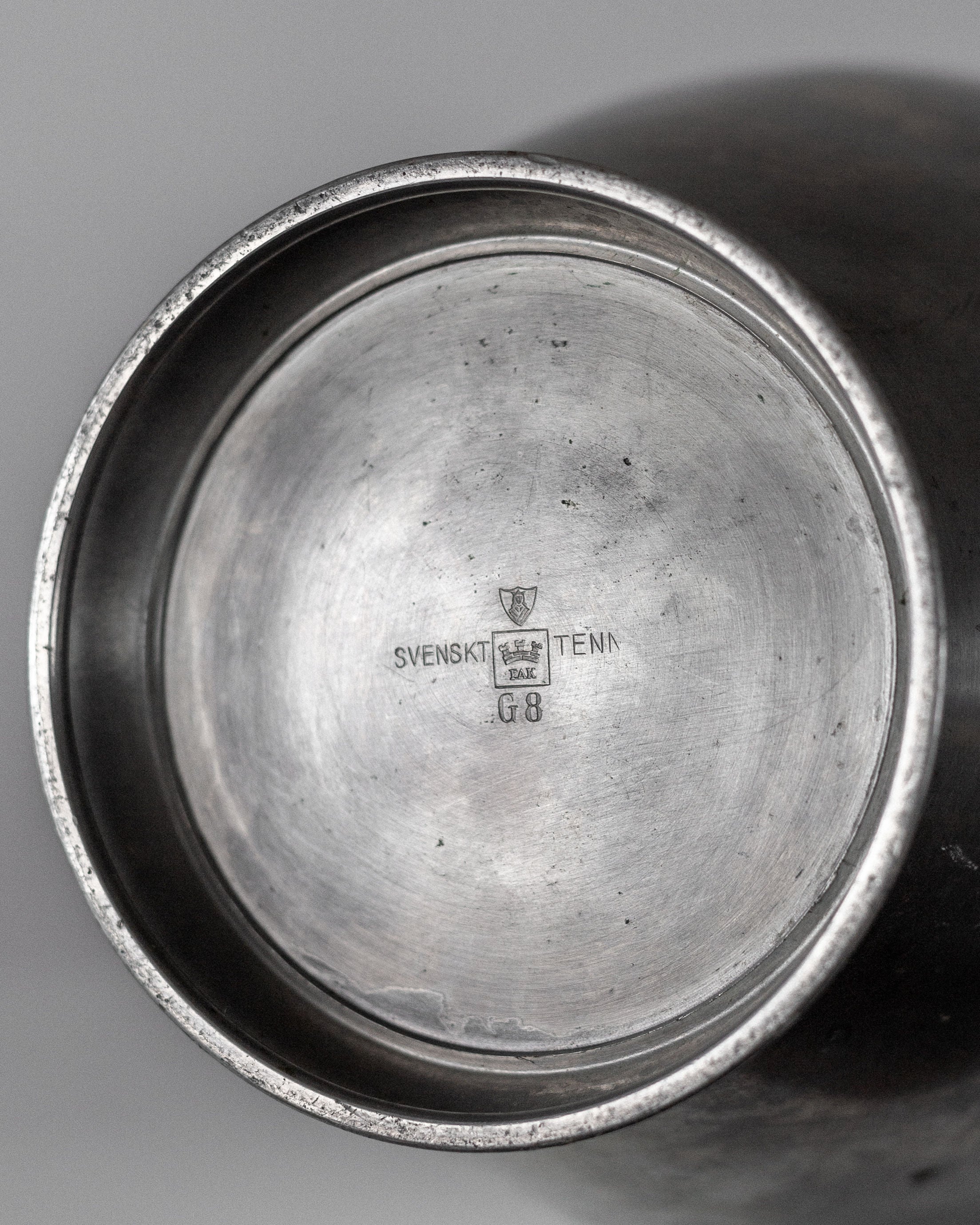 A close-up of the Large Modernist Metal Amphora by Spigel highlights its underside, displaying a maker's mark with a crest. "SVENSKT TENN" and "G8" are engraved in a brushed metal finish, reflecting Scandinavian influences and Rolf Engströmer's modernist style.