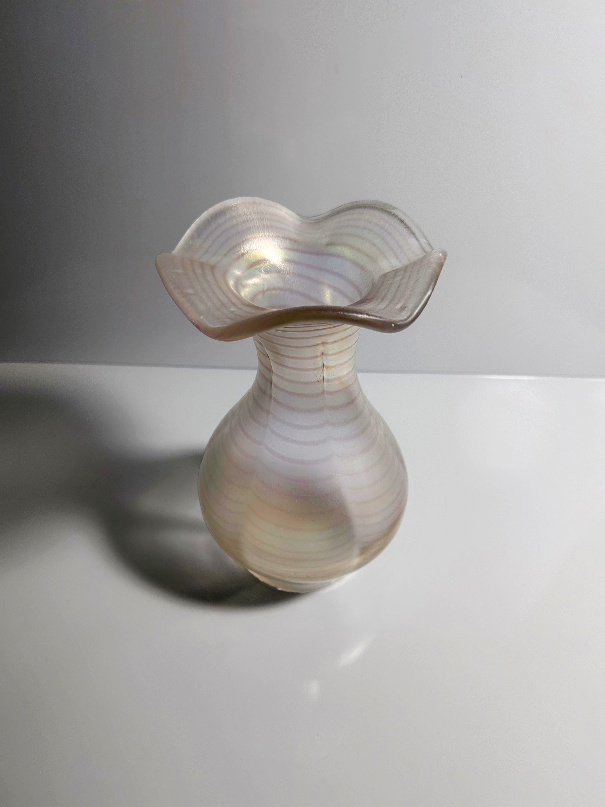 The Art Nouveau Wavy Vase by Dodo Vintage, featuring an iridescent finish and a wavy, flared rim with a bulbous base, reflects soft light. When set against a neutral background, its translucent and colorful stripes are reminiscent of Gallo’s enchanting designs.