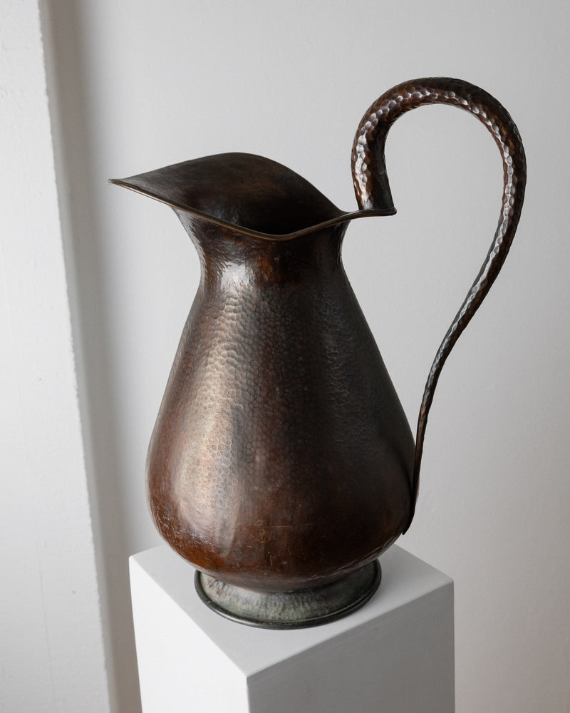 A Copper Handmade Jug/Vase by Bottega Jacobs from the 1950s, featuring a curved spout and handle, sits on a white pedestal against a plain wall. This Italian-crafted piece showcases fine craftsmanship with its textured surface, offering rustic and antique decor appeal.