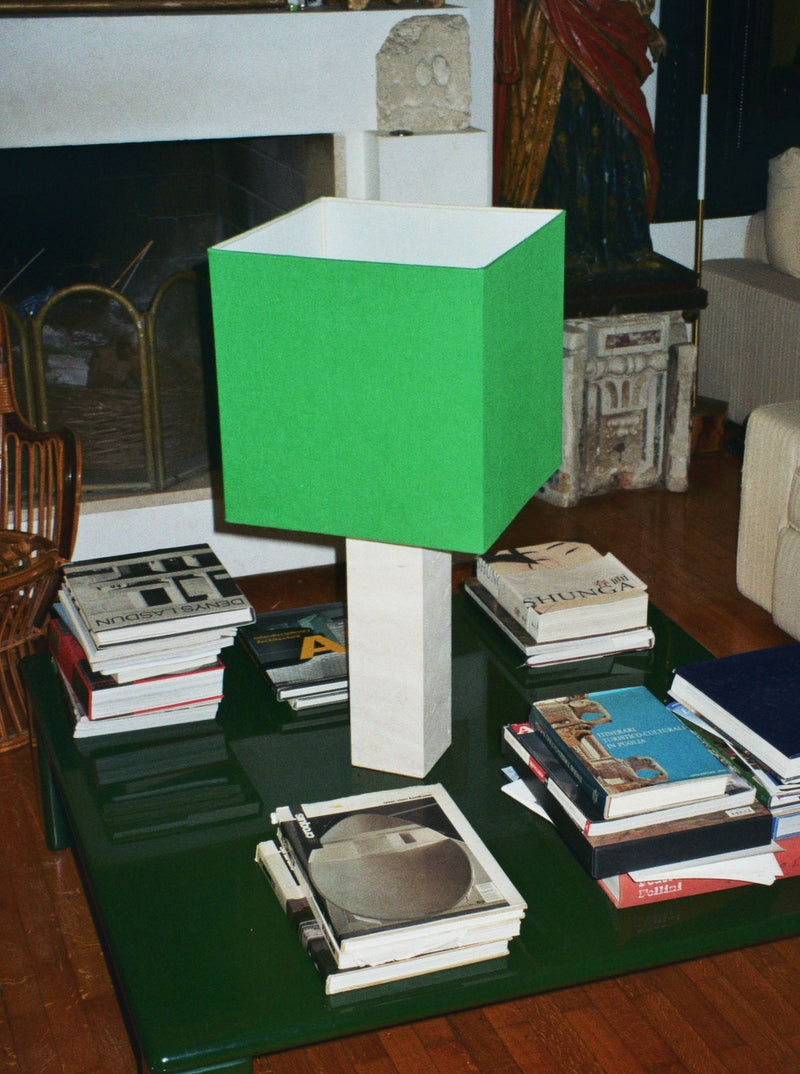 In a modern living room, a limited edition Roma I Lamp Squared in White Travertine and Green Linen from Nassi Lamps sits on a glass-top coffee table. The table is covered with stacks of books surrounding the lamp. In the background, there's a fireplace, wicker chair, and an upholstered sofa.
