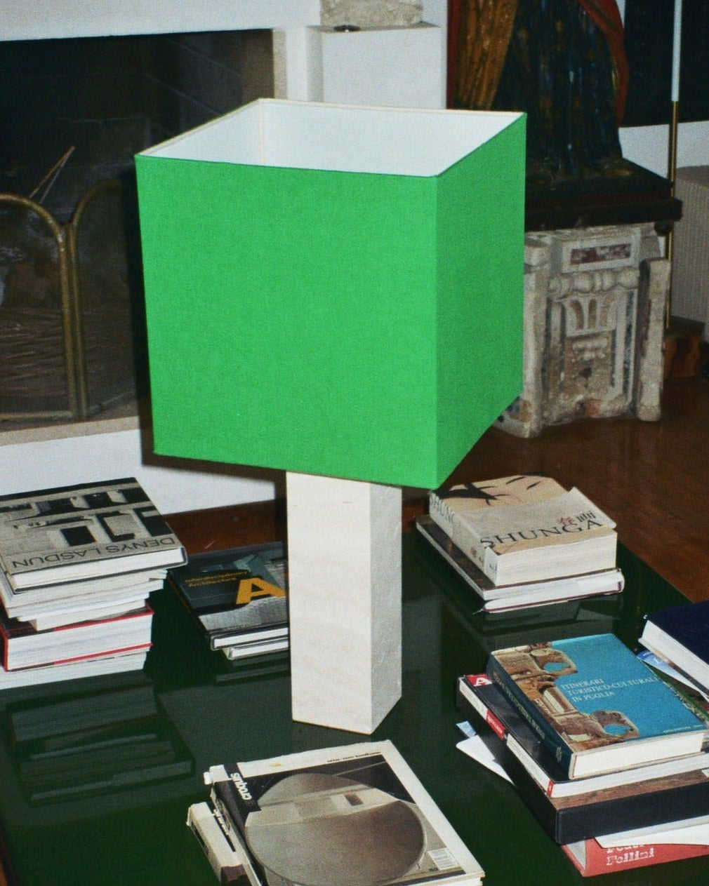 In a modern living room, a limited edition Roma I Lamp Squared in White Travertine and Green Linen from Nassi Lamps sits on a glass-top coffee table. The table is covered with stacks of books surrounding the lamp. In the background, there's a fireplace, wicker chair, and an upholstered sofa.
