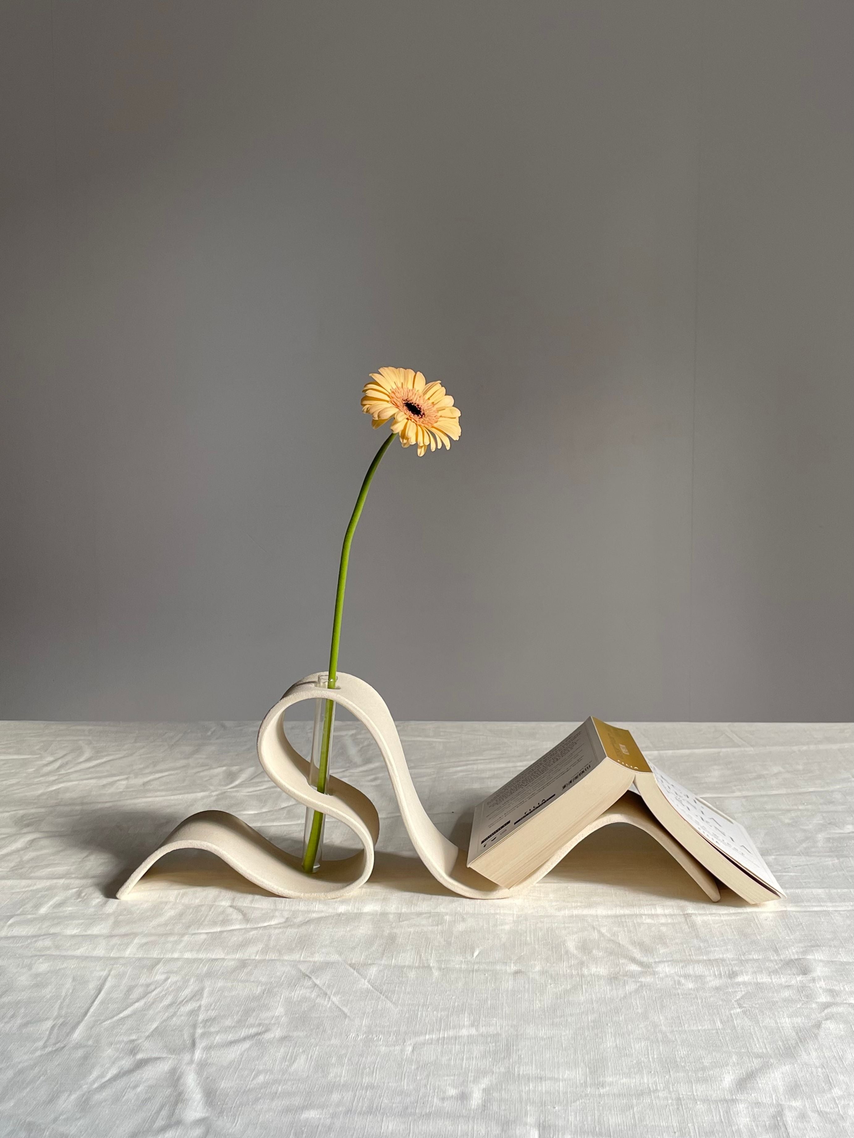 A single yellow flower in a LESS Ribbon Vase in Off White holds an open book on a light-colored, textured surface against a plain grey background. The vase's curves support both the flower and the book, creating an artistic and delicate arrangement reminiscent of an elegant book holder.