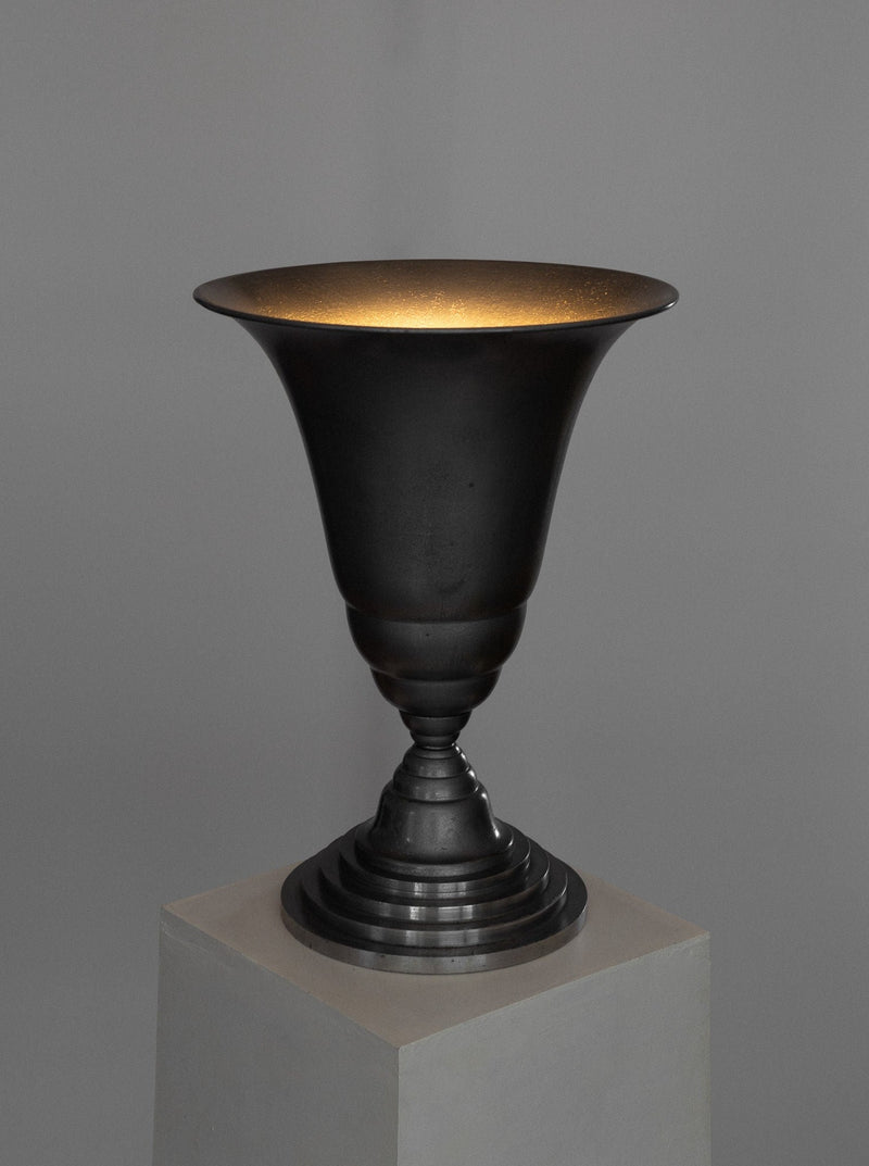 A dark, metallic Art Deco Torchière Table Lamp from France with a wide-brimmed and smooth, concave shape along with a textured base, displayed on a simple white pedestal against a gray background. The lighting creates a soft glow inside the lamp reminiscent of French Art Deco lighting, highlighting its polished surface and elegant design under the renowned brand Spigel.