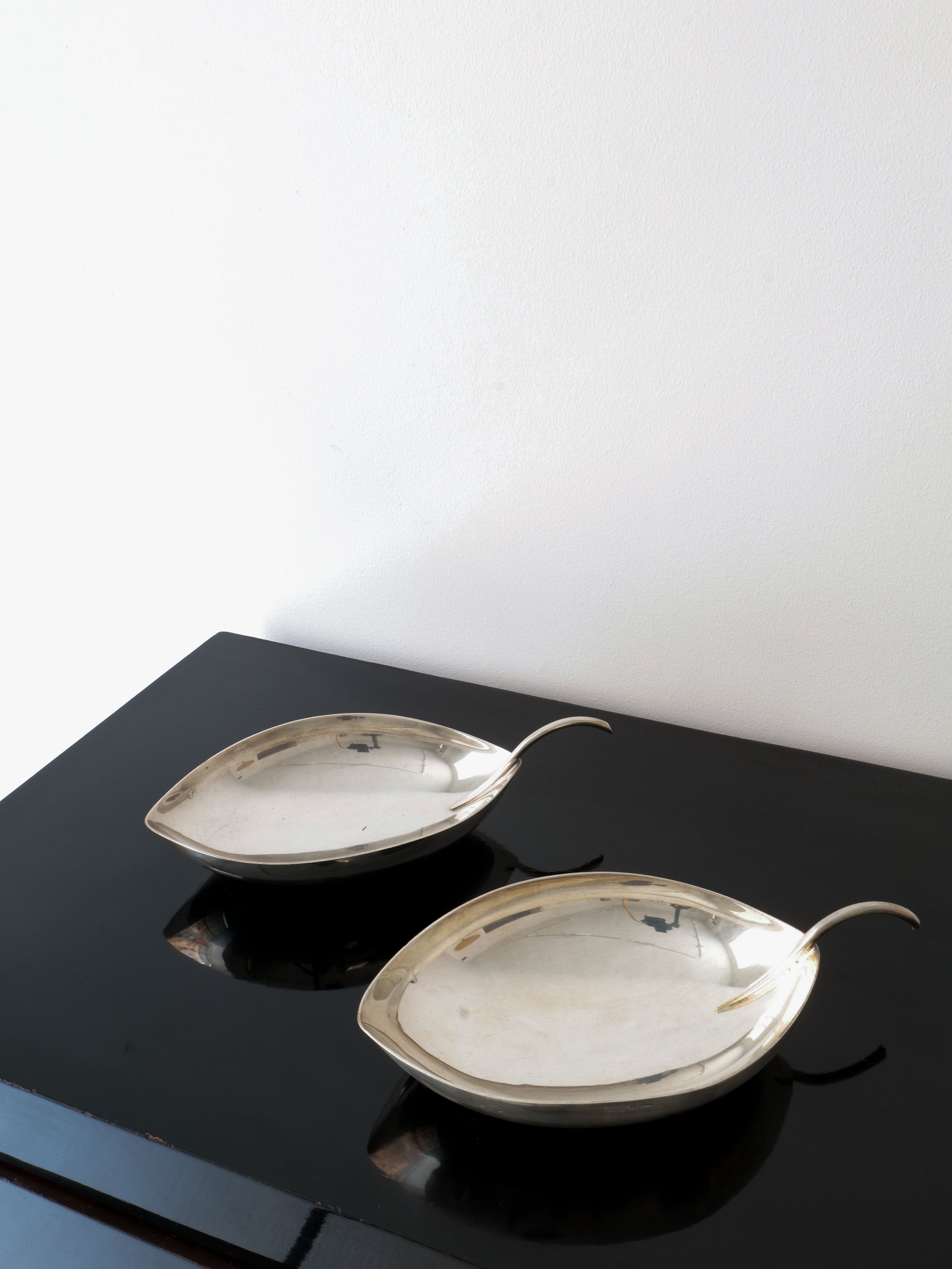 Two "Leaf Vide Poche" bowls by Christofle, designed by Lino Sabattini and from the Collection apart brand, rest elegantly on a sleek black surface against a plain white wall. The minimalist design exudes modern elegance.