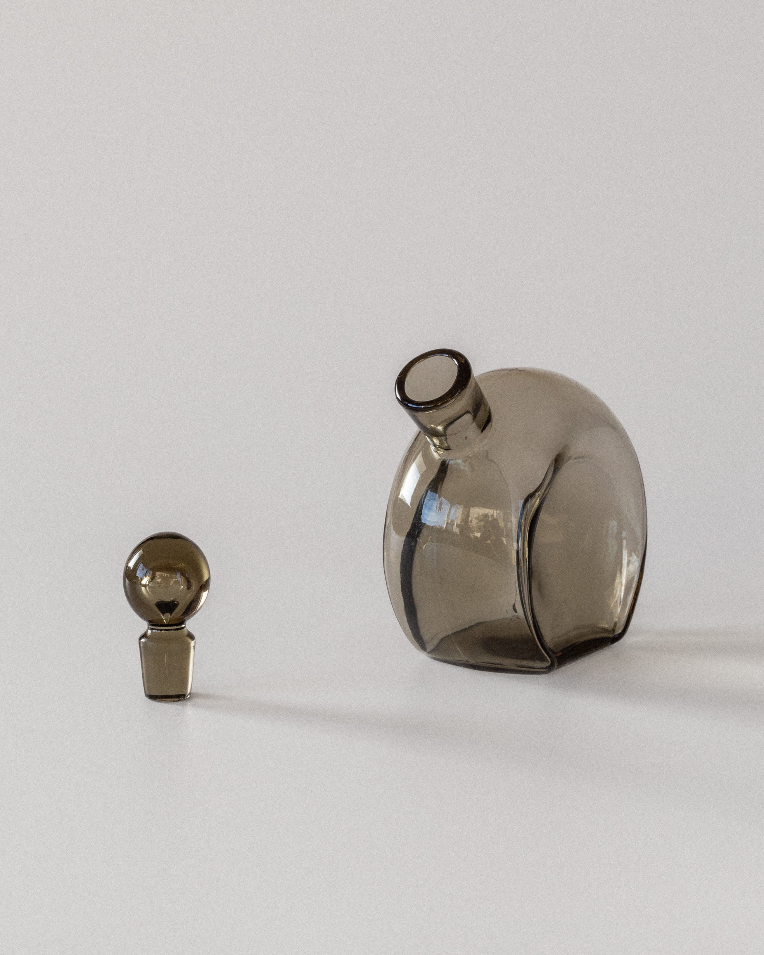 Two brown-toned glass objects on a light gray background: a round, tilted Glass Decanter by Harald Notini for Spigel, paired with a smaller spherical piece. Both showcase elegant Swedish modernism through their sophisticated smoky brown glass aesthetics.