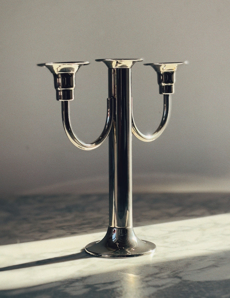 The Huerto 3 Arm Metal Candleholder, exhibiting an art deco vibe, stands on a marble surface. Sunlight casts its shadow, highlighting the sleek silver finish against a plain, light-colored wall.
