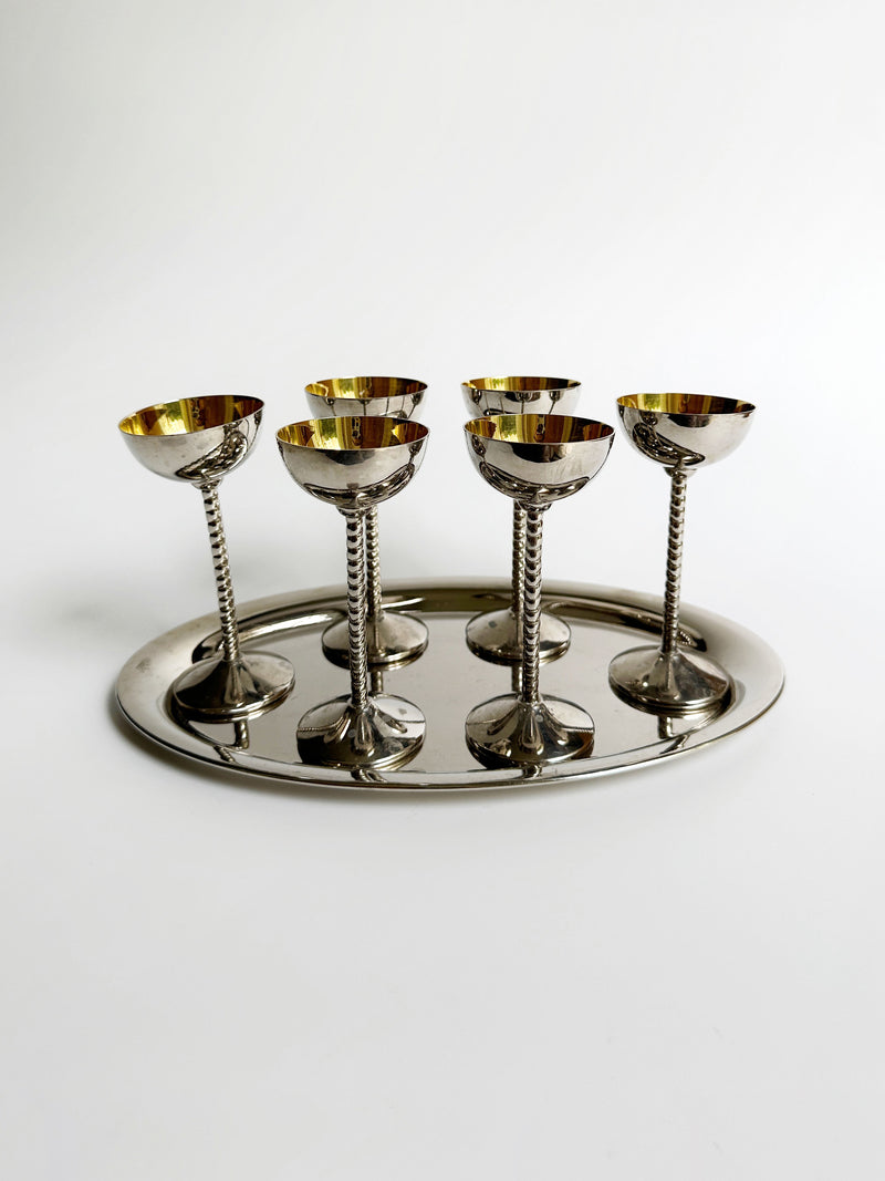 A set of six silver Art Nouveau liqueur goblets with twisted stems, from Dodo Vintage, arranged symmetrically on an oval silver tray. The polished and reflective goblets are standing upright with their open ends facing up against a plain white background.