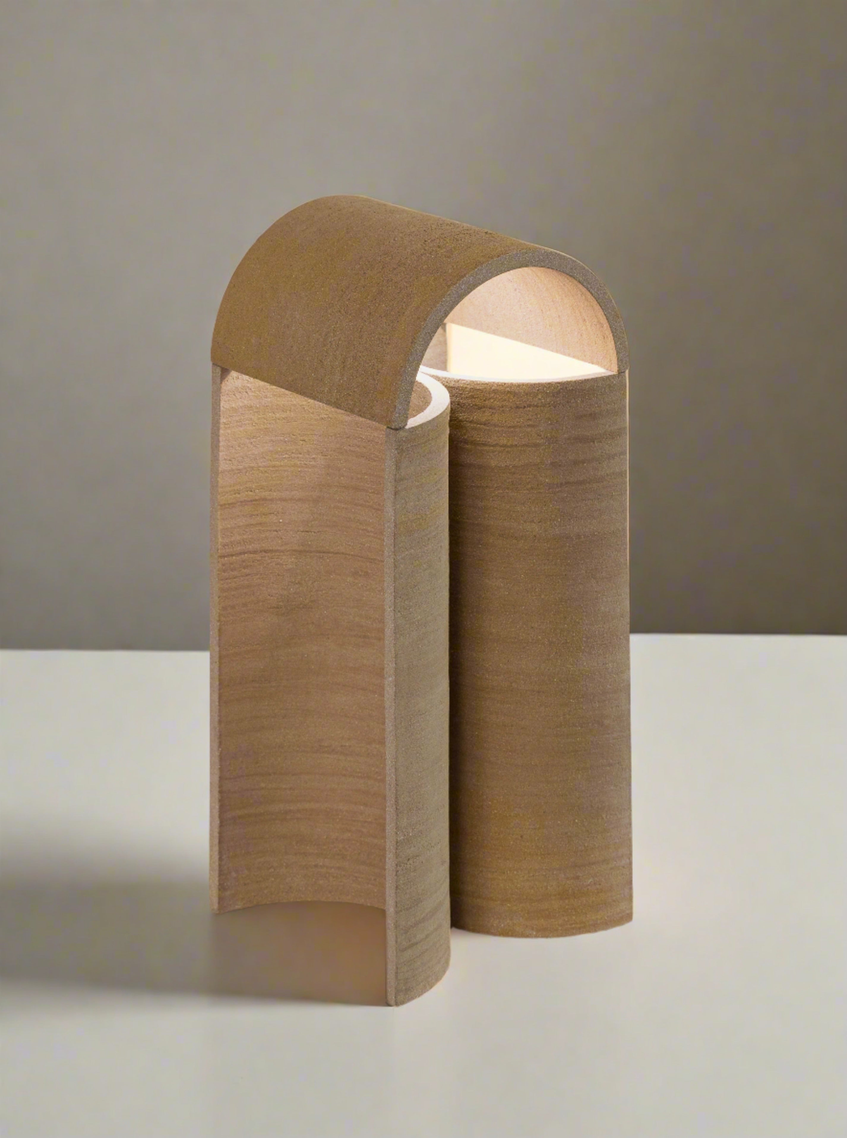 A modern, minimalist table lamp with a wooden outer shell is placed on a white surface. Its curved design partially encloses the light source, casting a warm glow. This sculptural lighting piece, the "Layers of..." Light II by Evelina Kudabaite, is complemented by a green plant leaf in the upper right corner, adding a natural element to the scene.