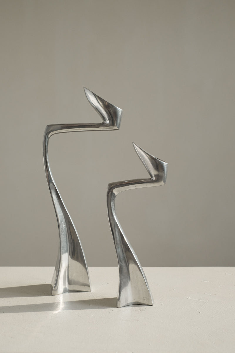 The Pair of Candleholders by Matthew Hilton 80's, from septembre studios, features two abstract, silver metallic sculptures with sleek, curved forms. They display elongated shapes resembling figures facing each other on a light surface against a plain background, captivating design enthusiasts.