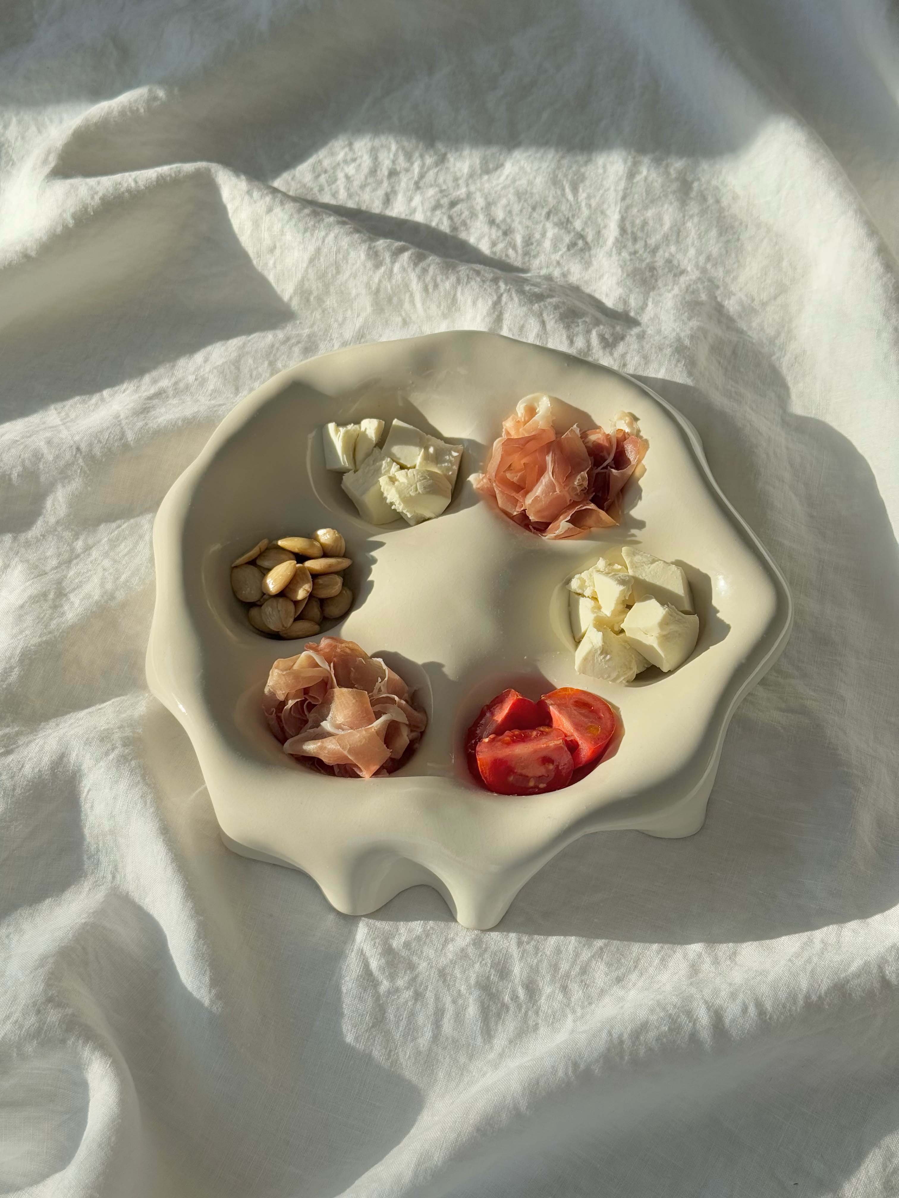 The Goobs Ceramics hand-made Ceramic Snack Plate, with its high gloss glaze, elegantly displays almonds, cheese cubes, prosciutto slices, and tomatoes on crumpled white fabric, capturing light and casting gentle shadows.