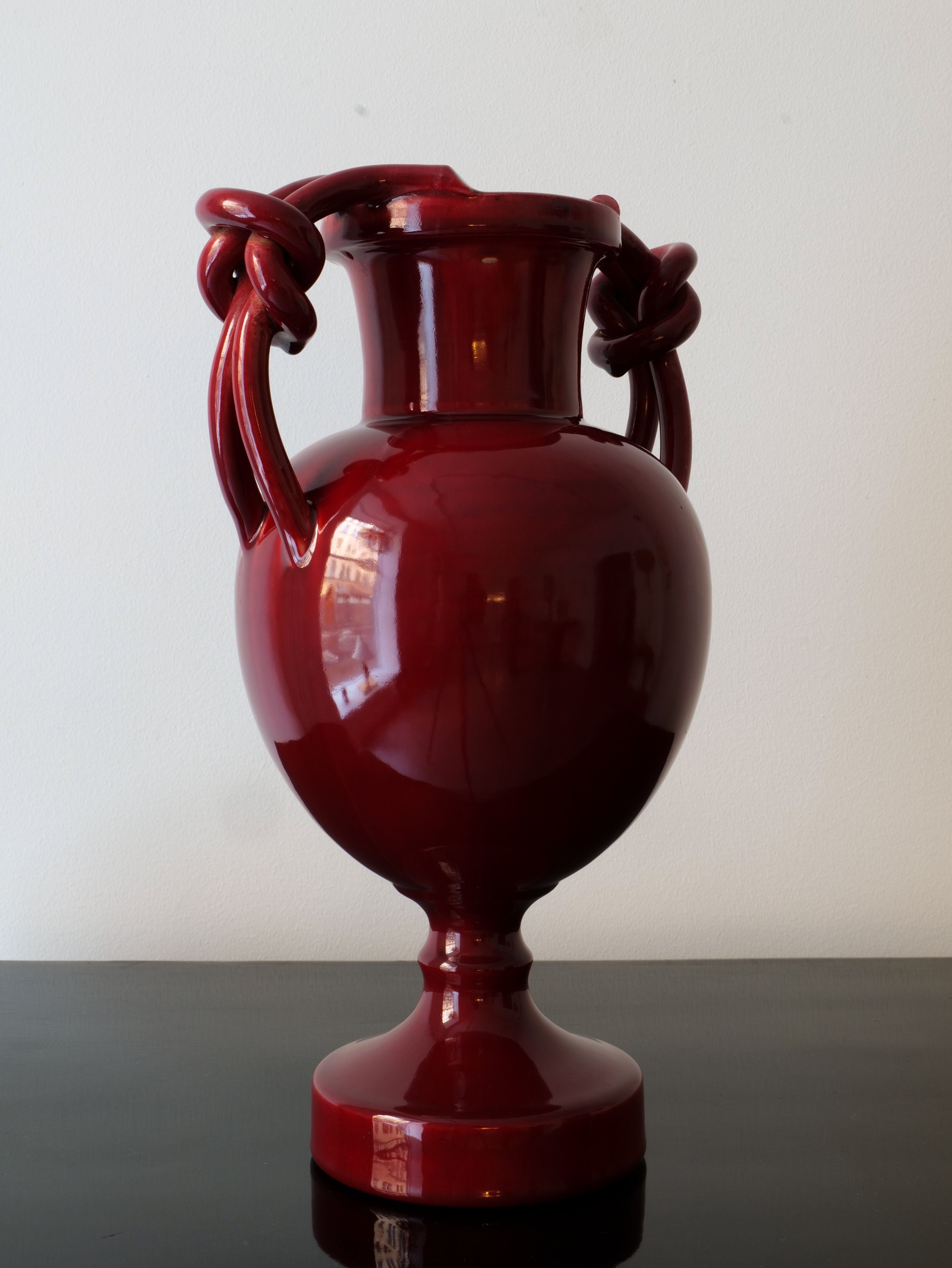 A glossy, dark red ceramic Baluster Vase Jérôme Massier by Collection apart, reminiscent of Vallauris ceramics, features two intricately knotted handles and rests on a polished black surface. The vase's reflective finish captures the light, emphasizing its smooth and elegant form against a plain white background.