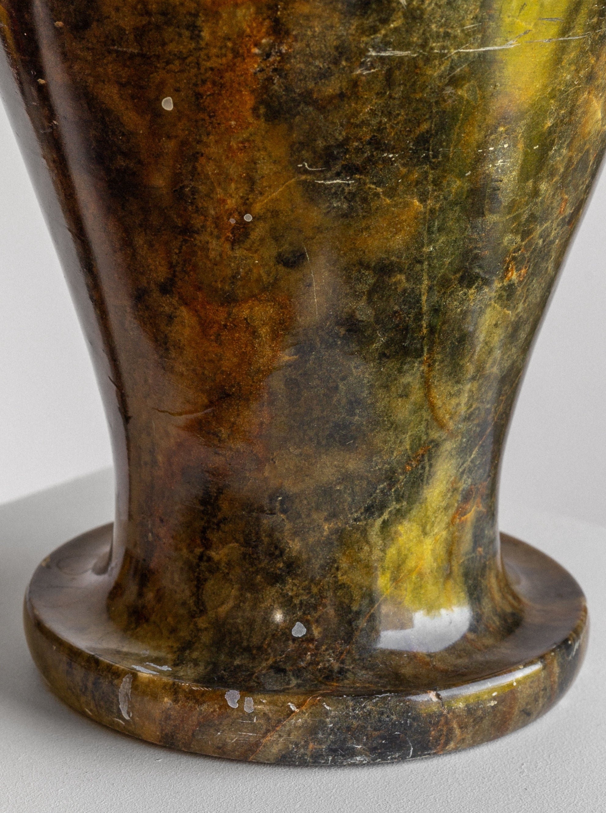 A close-up of the beautifully polished Large Neoclassical Stone Vase by Spigel, placed on a white pedestal. This stunning vase features a rich, marbled texture with earthy tones of brown, yellow, and green. Light reflections highlight the smooth surface, showcasing its intricate patterns and natural variations.