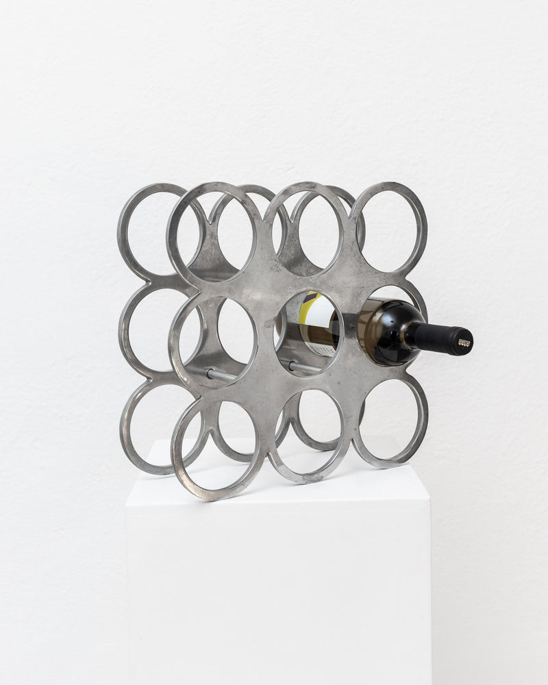 A Vintage metal wine rack by Bottega Jacobs, featuring a geometric Space Age design from the 1970s, is displayed on a white pedestal. This silver piece, capable of holding nine bottles, is set against a plain white wall to evoke the sleek style of its era.