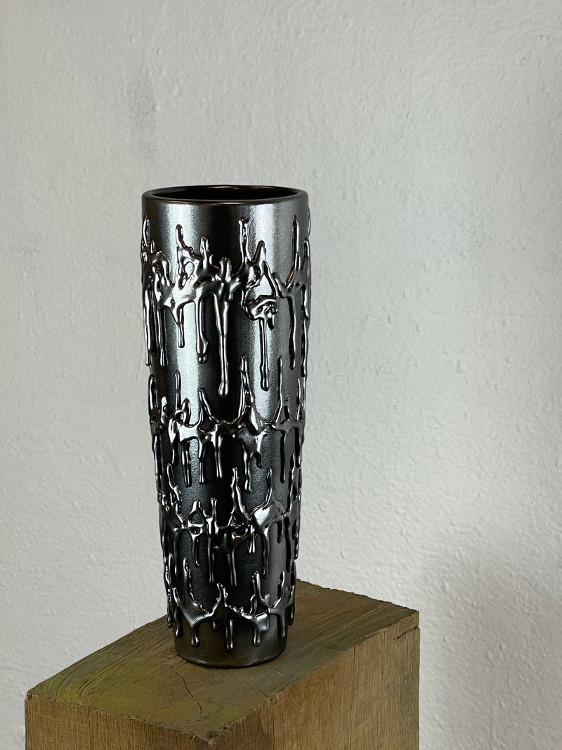 The Almost Chrome Vase .001 by John Nordenstein is a tall, dark handmade ceramic vase featuring a shiny metallic finish and raised abstract figures. This sculptural chrome piece stands on a wooden block against a plain wall.