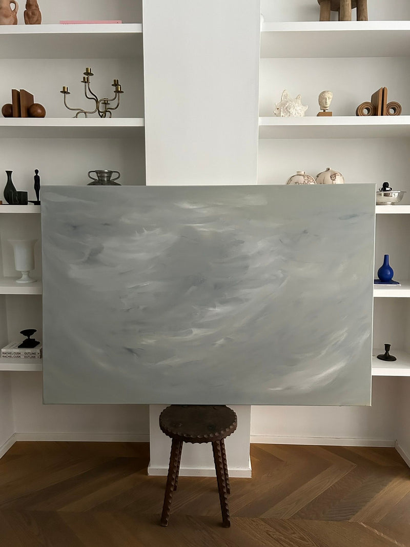 The expansive abstract artwork "Veil of Tranquility" by Mattias Storm, characterized by swirling gray and white patterns, rests on a compact wooden easel. White shelves in the background display an array of decorative items, including vases, candles, and sculptures, all set against a wooden floor.
