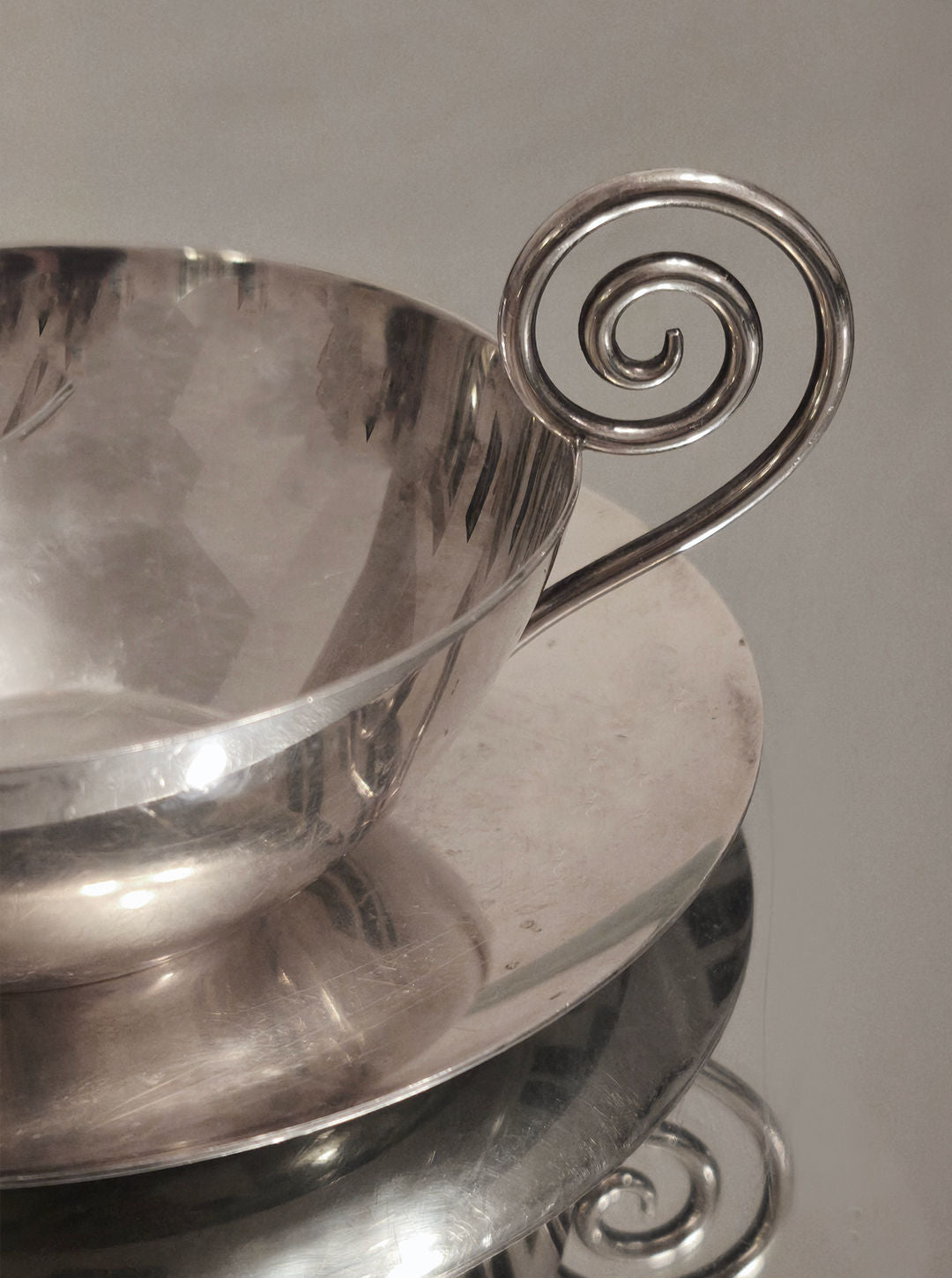 A close-up of a luxury collectible, the Les Objoies Spiral Christofle Cup with its decorative spiral handle, sits elegantly on a reflective silver saucer, perfectly mirrored in the pristine surface beneath.