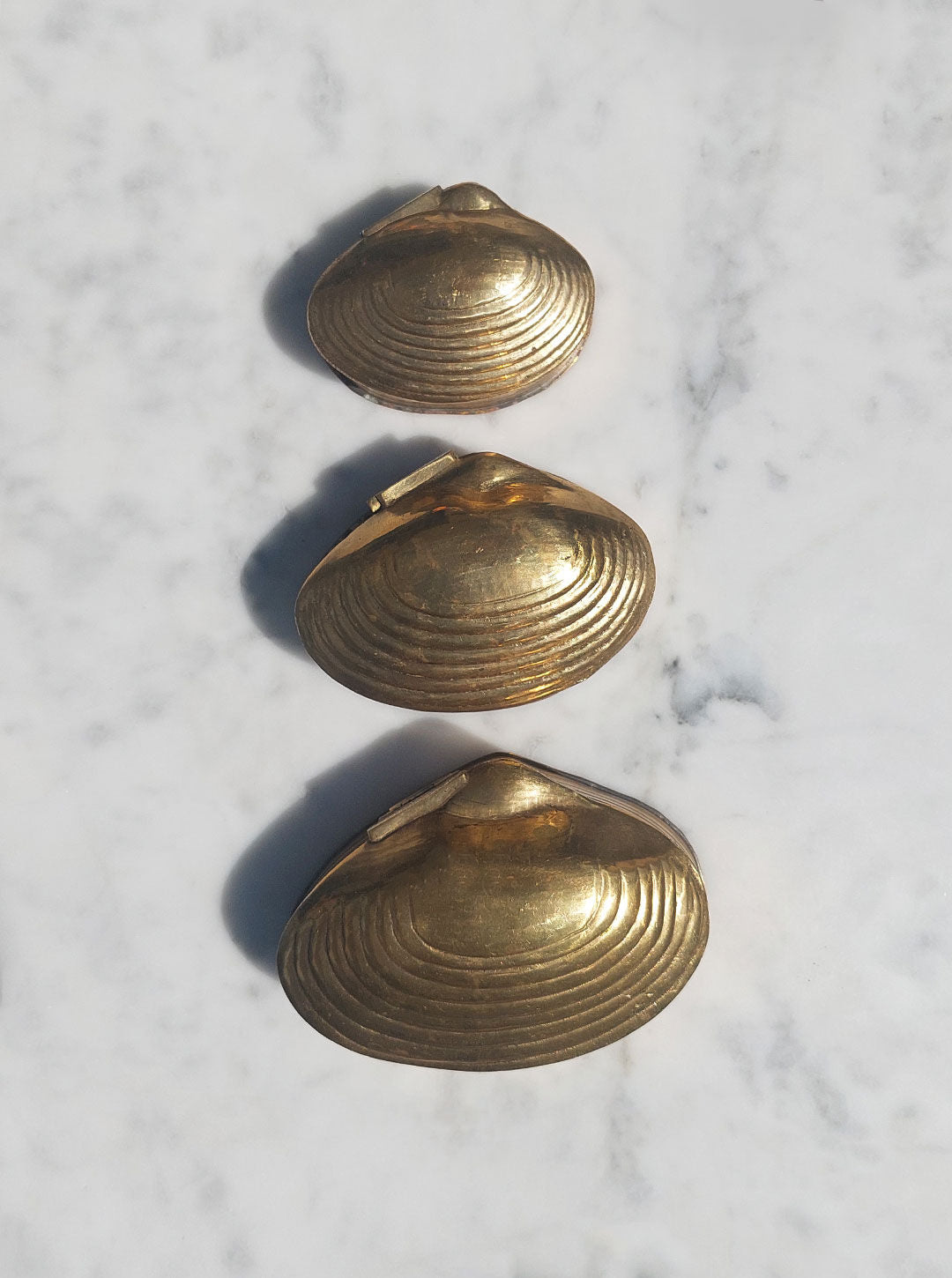 Three gold clam shell-shaped objects of varying sizes are arranged vertically on a white marble surface. The objects, with their brass gold finish, have a metallic sheen and are possibly decorative items or nesting boxes. The arrangement forms a gradation from the smallest at the top to the largest at the bottom. This is the Set of 3 Nesting Shell Boxes by Les Objoies.