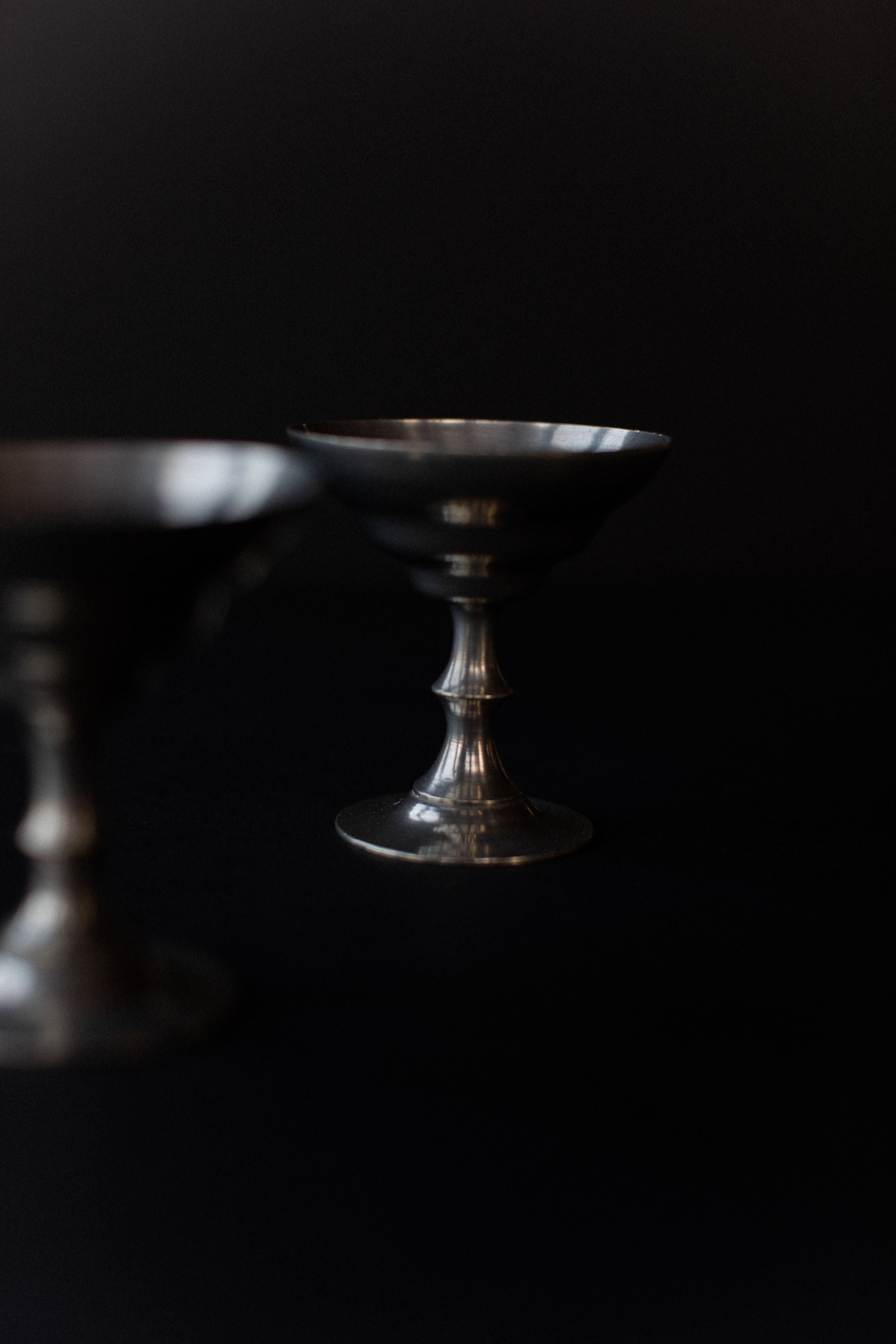 In a dimly lit image, two dark metallic goblets from Out For Lunch's Ice Cream Cups collection stand against a black background. The focus is on the back goblet with its vintage geometric design, while the foreground cup is slightly blurred.