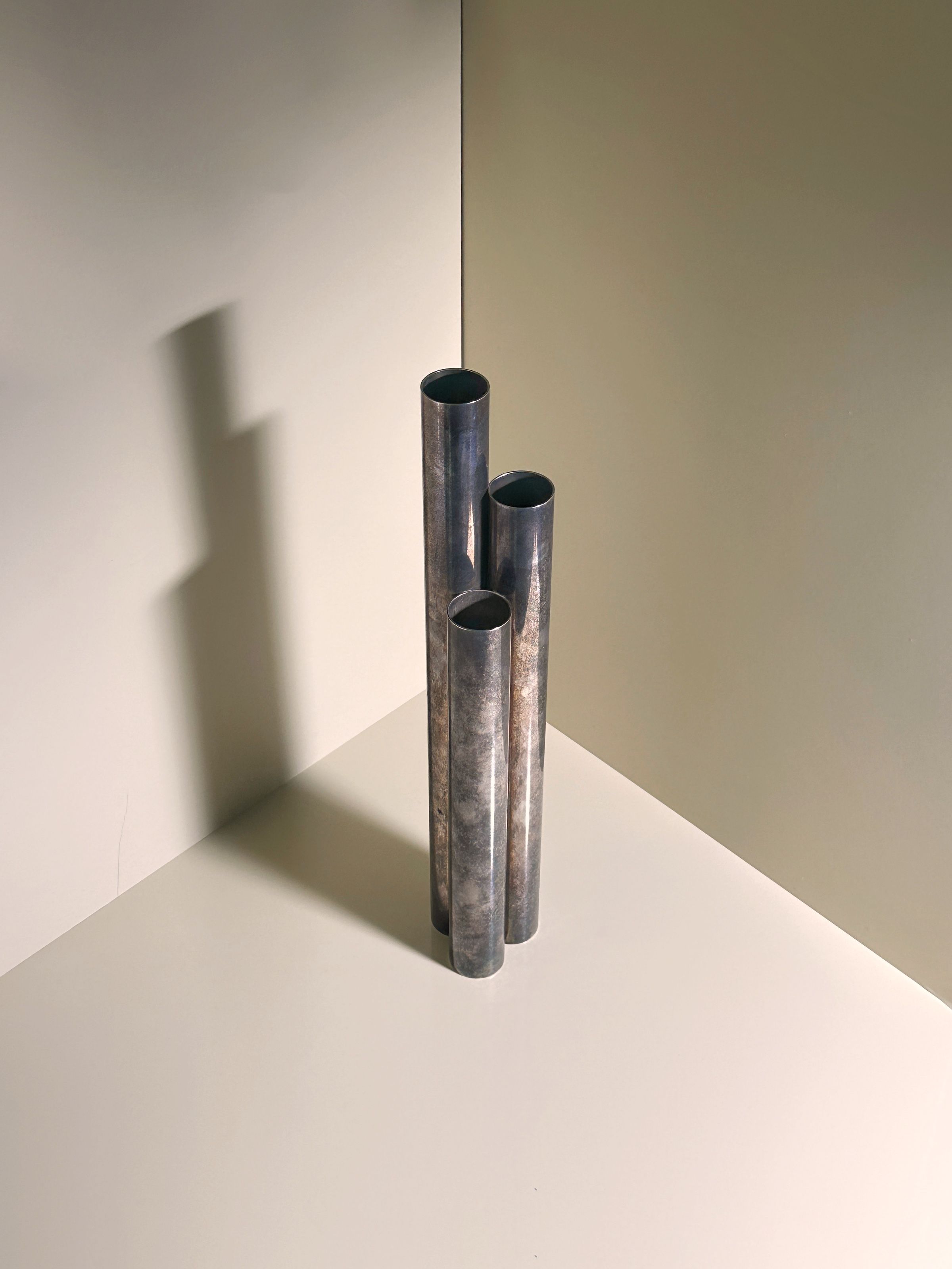 The Silver-plated Tall Brutalist Vase by Dodo Vintage, Gio Ponti Italia 1960s, features three minimalist cylindrical sections casting soft shadows on a light surface in a corner.