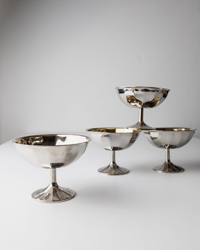 Four Bottega Jacobs Champagne Coupes from Italy in the 1970s, with silver-plated elegance, are set on a white surface—three forming a pyramid on the right and one standing alone on the left. The plain gray wall in the background enhances their timeless allure.